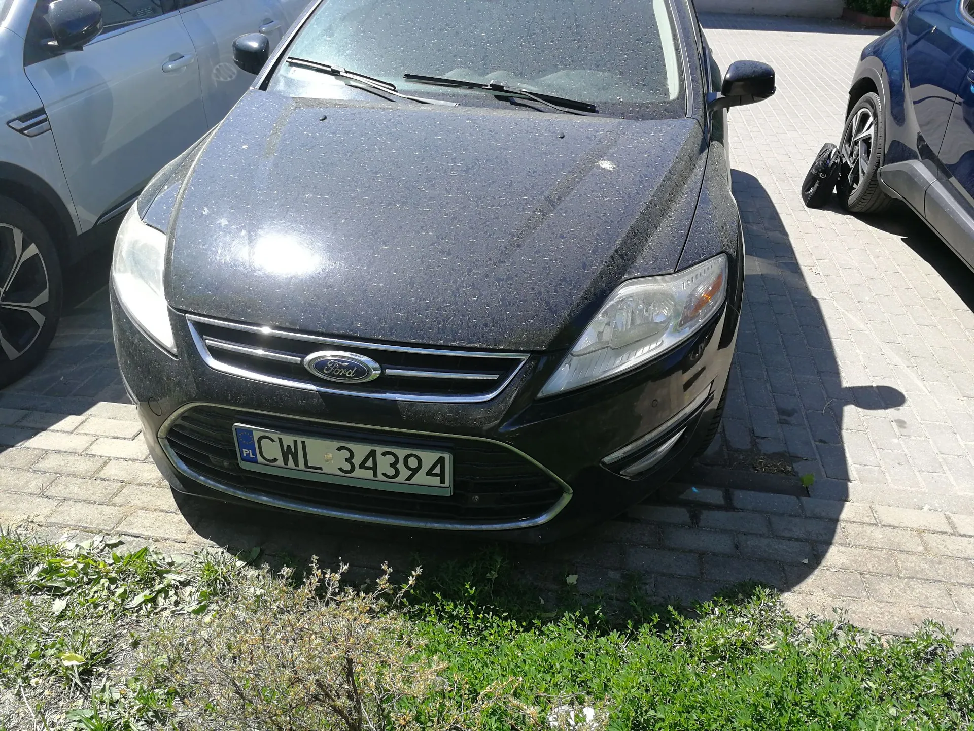 Czarny samochód marki Ford zaparkowany na kostce brukowej. Karoseria samochodu jest pokryta grubą warstwą kurzu i pyłu. Na przodzie widoczna jest polska tablica rejestracyjna o numerze CWL 34394. Po lewej stronie fragment białego samochodu, a po prawej fragment ciemnego pojazdu.