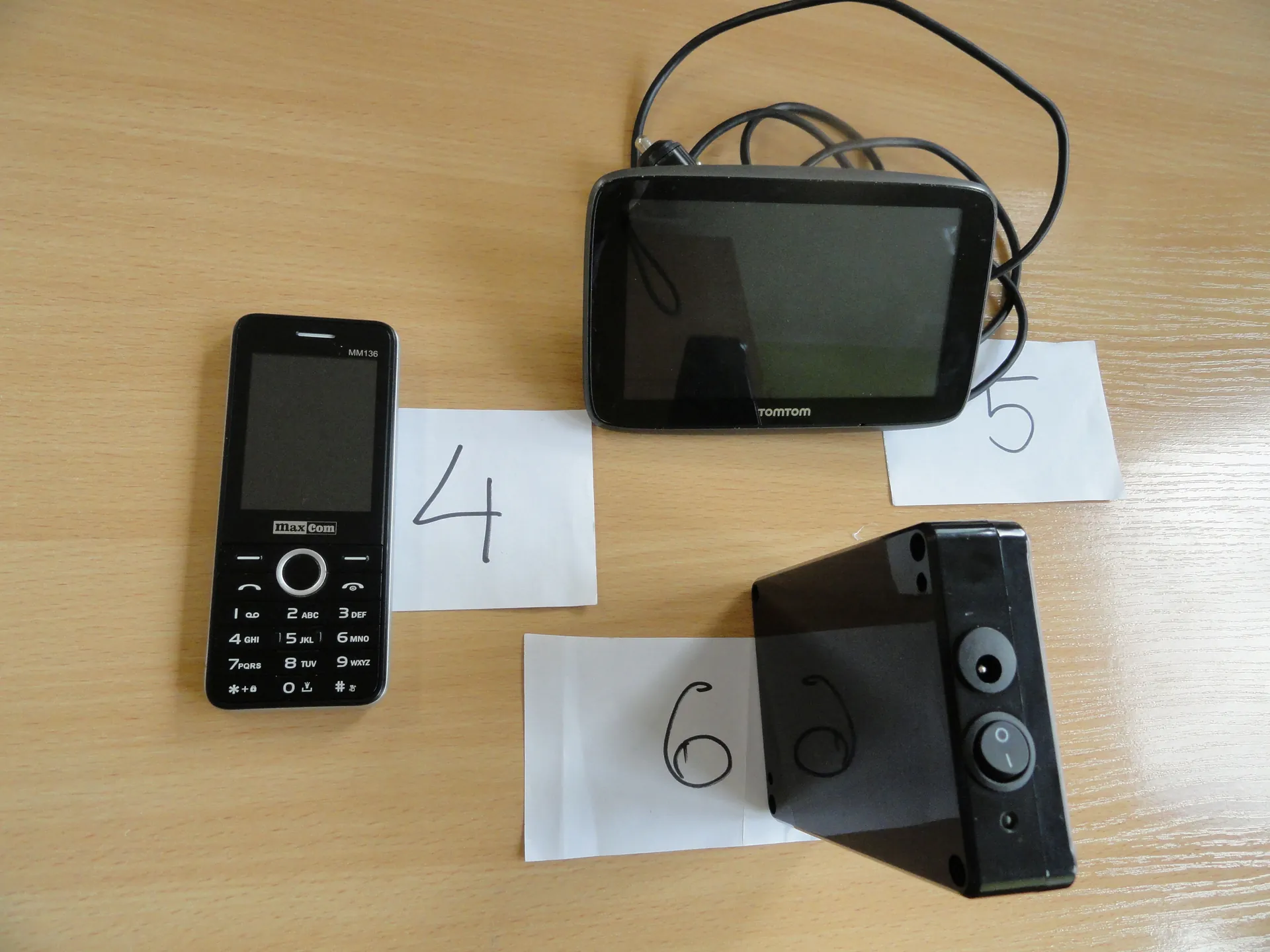 Trzy urządzenia elektroniczne na drewnianym stole: czarny telefon komórkowy marki MaxCom model MM136 z klawiaturą, nawigacja TomTom z ekranem i przewodami oraz czarny aparat lub urządzenie z dwoma przyciskami. Obok każdego urządzenia kartka z napisaną cyfrą: 4 pod telefonem, 5 pod nawigacją, 6 pod trzecim urządzeniem.