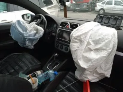 Wnętrze samochodu z rozwiniętymi poduszkami powietrznymi: niebieską po stronie kierowcy i białą po stronie pasażera. Widoczne są kierownica, deska rozdzielcza z przyciskami, dźwignia zmiany biegów oraz przedmioty na siedzeniu pasażera: butelka wody i kubek z słomką. Przez przednią szybę widać zaparkowane pojazdy i fragment budynku.