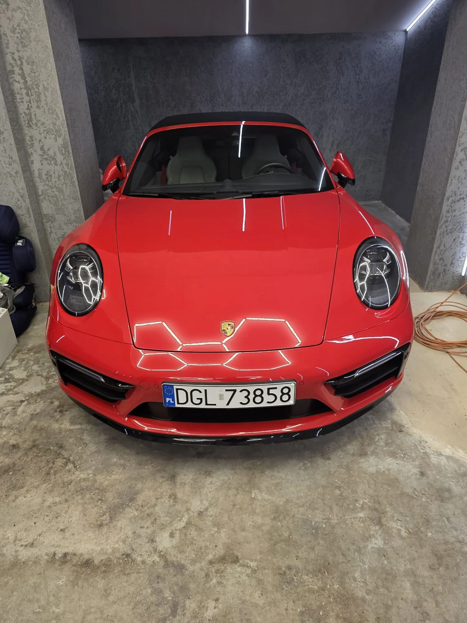 Czerwony samochód sportowy Porsche 911 Cabriolet z czarnym dachem, widok z przodu. Na masce widoczny emblemat marki. Tablica rejestracyjna PL DGL 73858. Wnętrze z jasnymi fotelami. Samochód znajduje się w pomieszczeniu z betonowymi ścianami i podłogą. Po lewej stronie widoczne są niebieskie torby, po prawej pomarańczowe przewody. Oświetlenie sufitowe tworzy refleksy na karoserii.