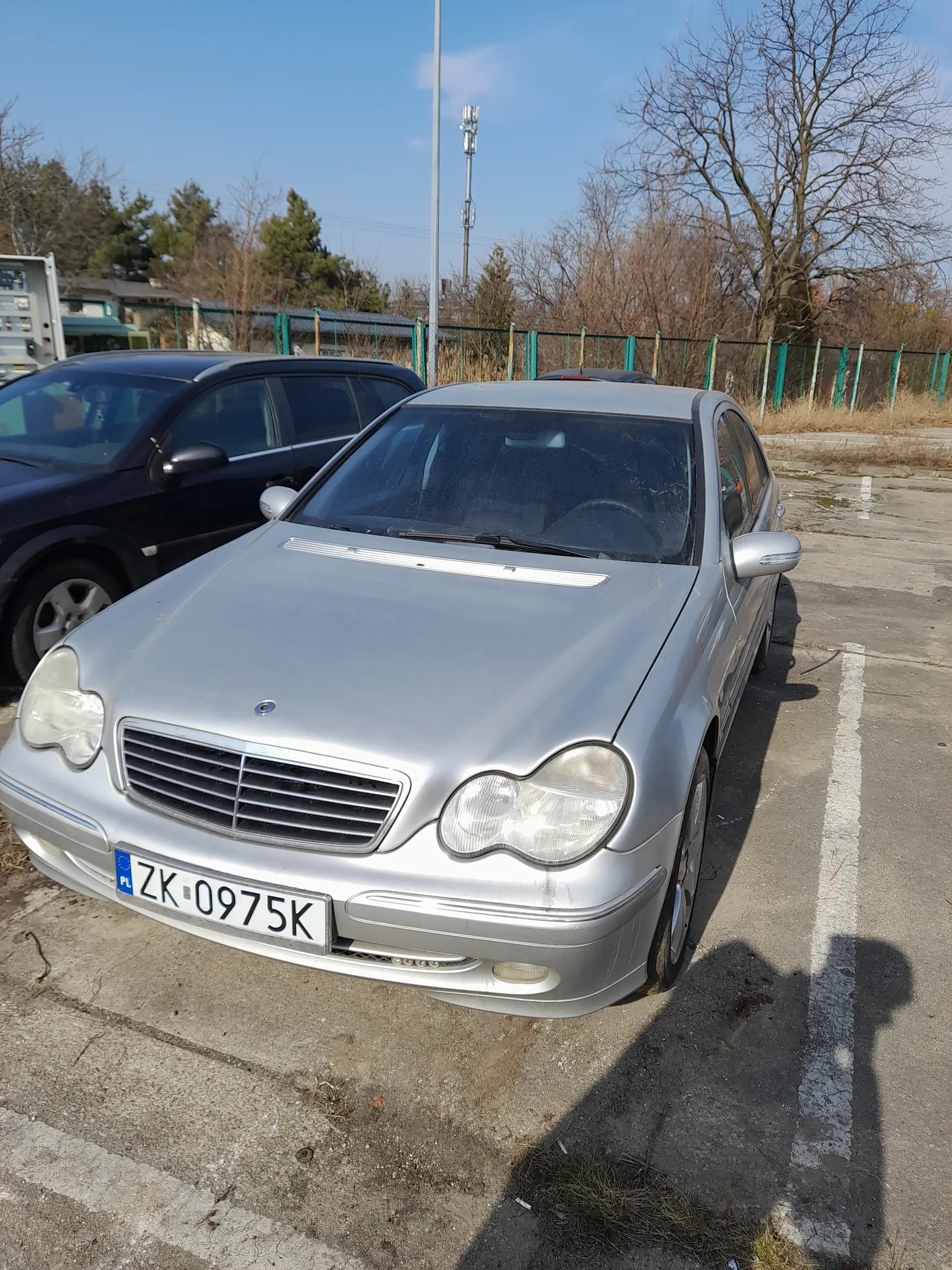 Srebrny samochód osobowy z tablicą rejestracyjną ZK-0975K zaparkowany na poboczu drogi. Obok niego stoi ciemny samochód. W tle widać zielone słupki ogrodzenia, drzewa bez liści, jasne niebo oraz cień osoby robiącej zdjęcie.