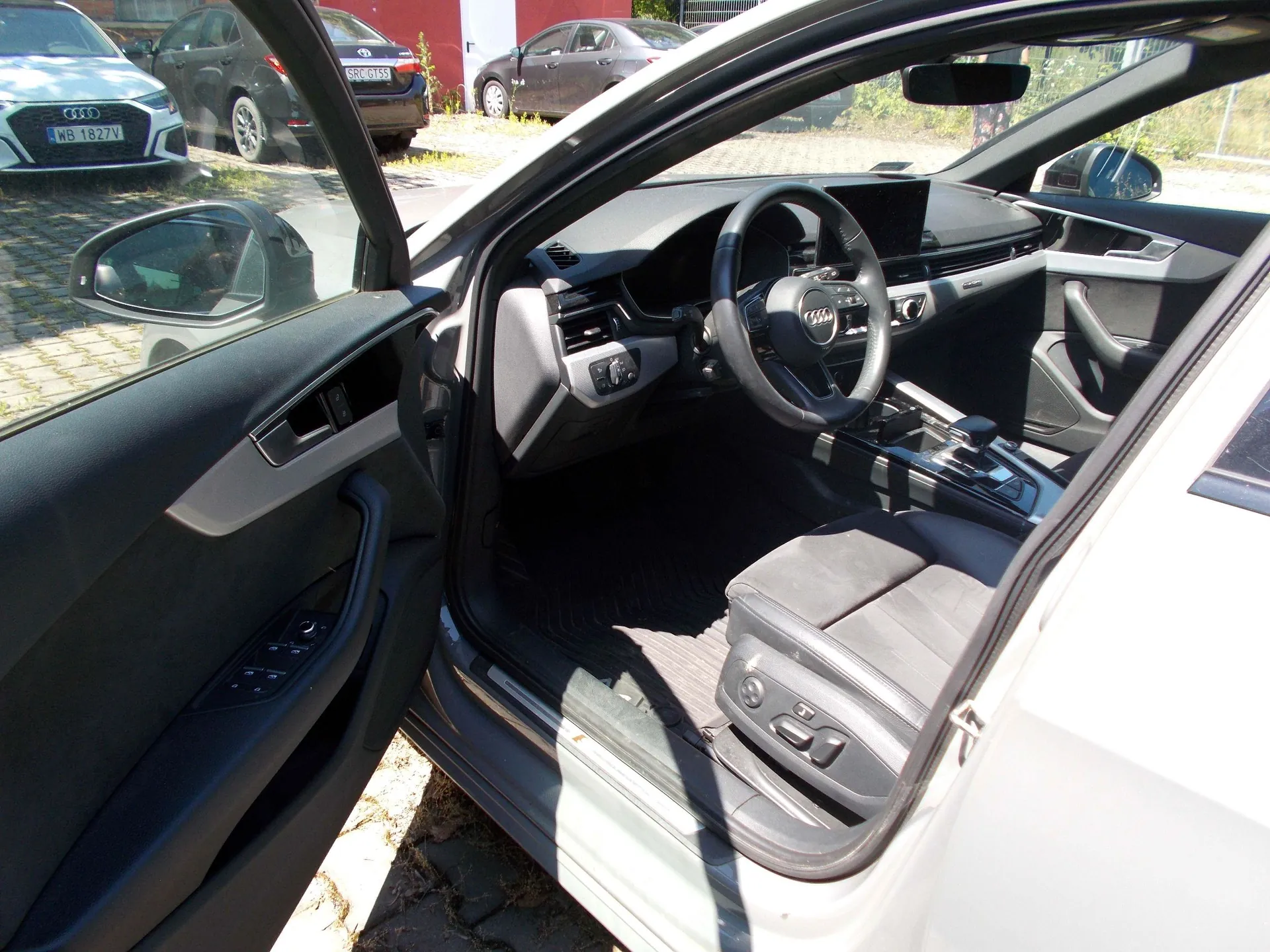 Widok z otwartych drzwi kierowcy do wnętrza nowoczesnego samochodu marki Audi. Widoczna jest czarna tapicerka, kierownica z logo Audi, deska rozdzielcza oraz konsola środkowa. W tle, na zewnątrz, zaparkowane są inne samochody, w tym białe Audi z tablicą rejestracyjną WB 1827V oraz ciemny samochód z tablicą CRC 6T53.