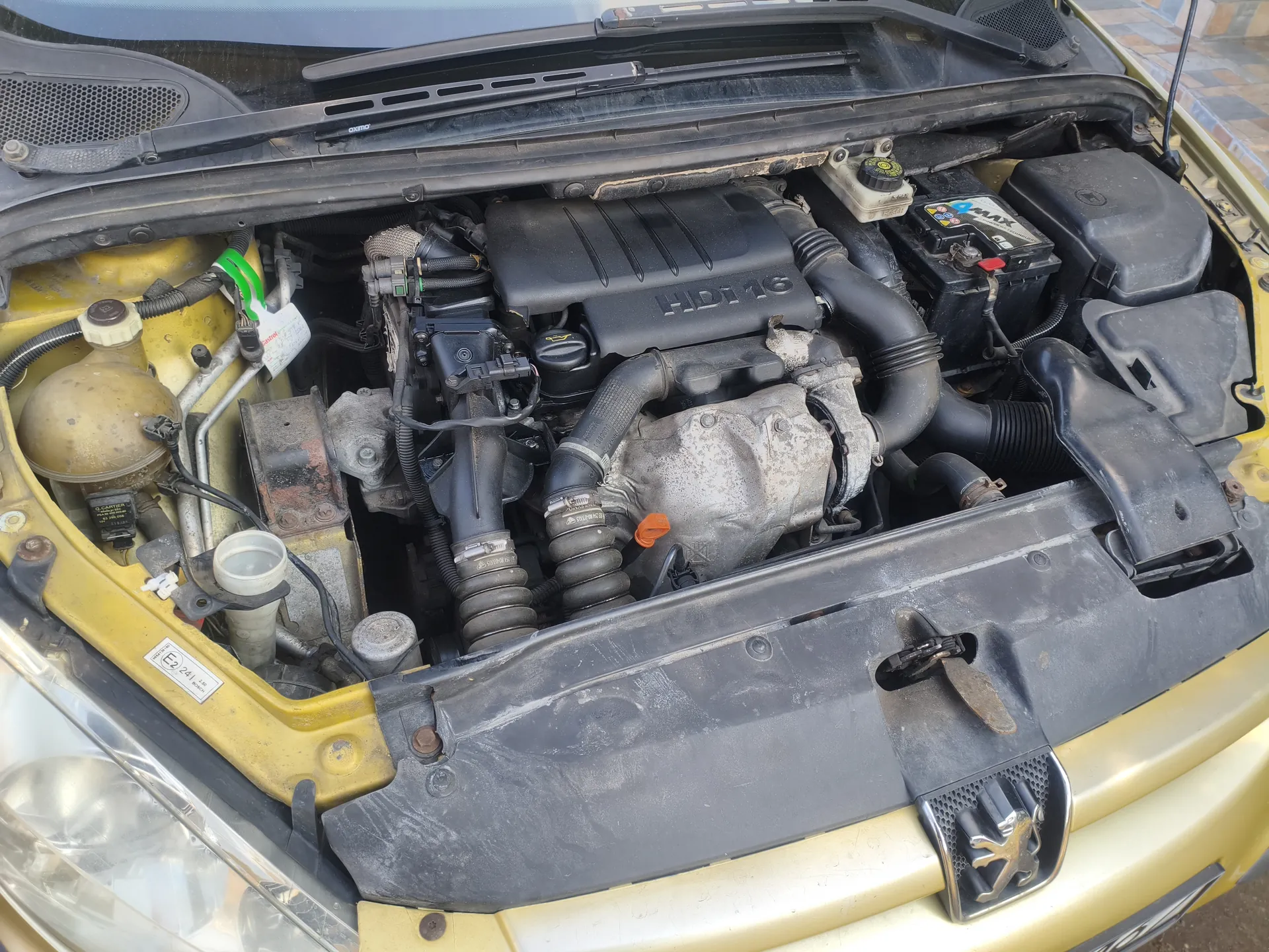 Widok otwartej tylnej części silnika samochodu marki Peugeot. Silnik oznaczony napisem HDi 16. Widoczne elementy: żółta karoseria, czarna pokrywa silnika, akumulator z etykietą, zbiornik płynu chłodniczego, przewody i rury. Na przedniej części nadwozia umieszczony jest emblemat Peugeot w postaci lwa. W lewym dolnym rogu widoczna tabliczka z oznaczeniem E2 24.