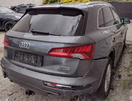 Tylny widok szarego samochodu marki Audi z widocznymi uszkodzeniami karoserii po prawej stronie tylnego zderzaka i błotnika. Na klapie bagażnika znajduje się logo Audi oraz napis TFSI quattro. Samochód nie posiada tablic rejestracyjnych, widoczny jest jedynie czarny uchwyt na tablicę.