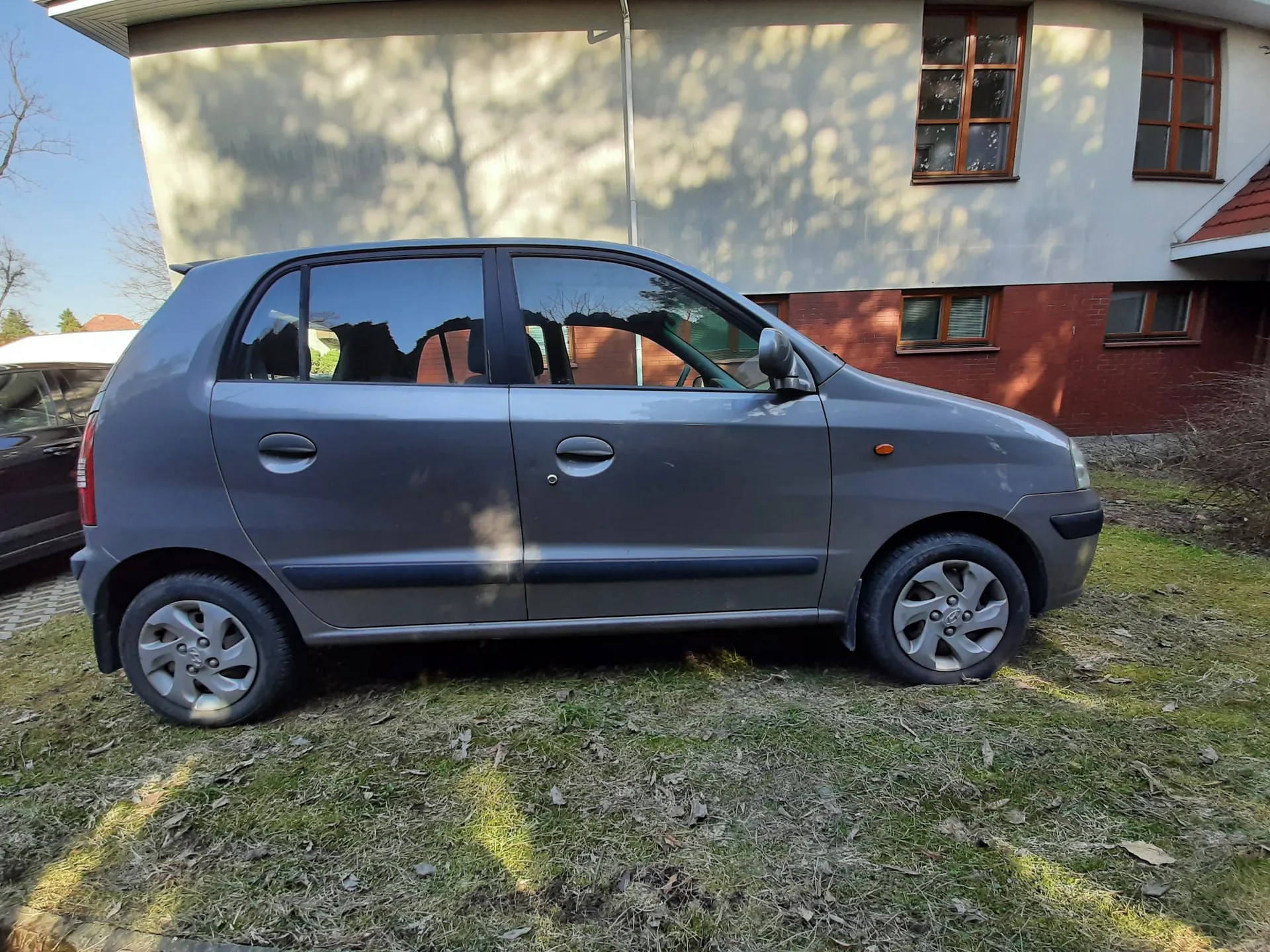 Szary samochód osobowy typu hatchback zaparkowany na trawie przed budynkiem. Budynek ma białą i czerwoną ceglaną elewację z oknami o brązowych ramach. W tle widoczne są drzewa i fragment innego pojazdu. Trawa jest częściowo sucha z rozsypanymi liśćmi.