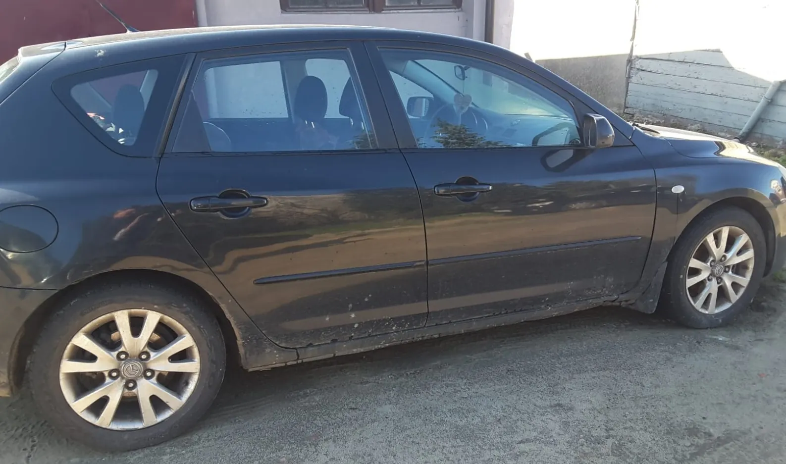 Ciemnoszary samochód osobowy typu hatchback zaparkowany na betonowej powierzchni. Pojazd ma cztery drzwi, srebrne felgi i zabrudzone boki. W tle widoczny jest budynek z czerwoną ścianą, oknem oraz drewnianą konstrukcją po prawej stronie. Słońce oświetla scenę, tworząc odbicia na karoserii.