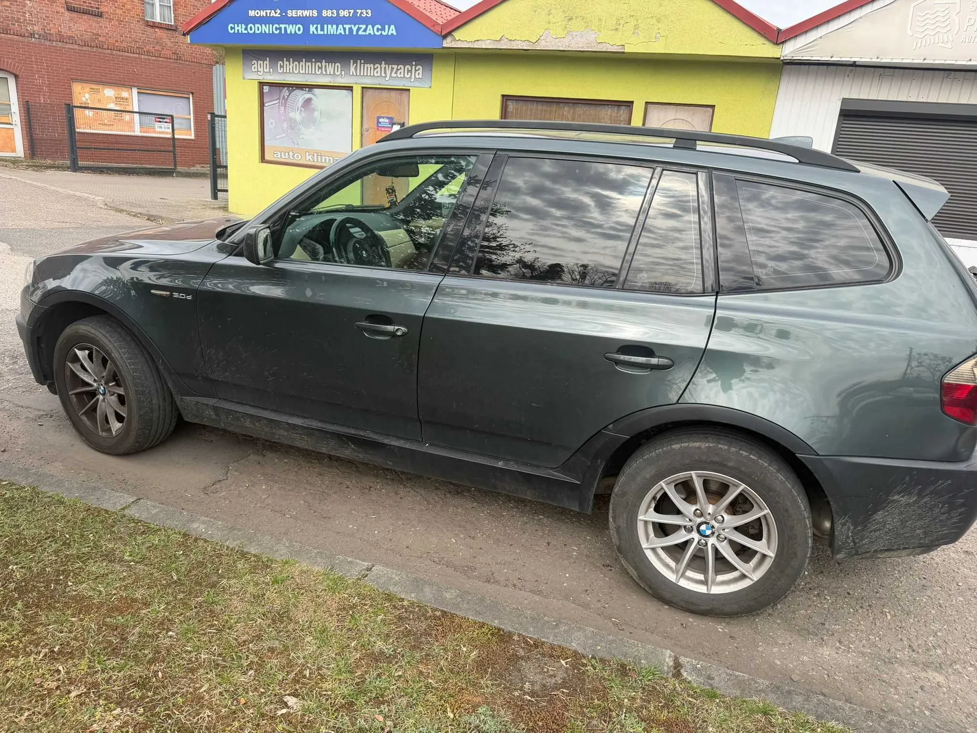 Ciemnozielony SUV marki BMW zaparkowany na poboczu. Samochód jest zabrudzony, widoczne są felgi z logo BMW. W tle żółty budynek z niebieskim napisem "MONTAŻ - SERWIS 883 967 733 CHŁODNICTWO KLIMATYZACJA" oraz szarym napisem "agd, chłodnictwo, klimatyzacja". Częściowo widoczny napis "auto klim" na szybie. Obok żółtego budynku ceglany budynek z ogrodzeniem i trawnik na pierwszym planie.