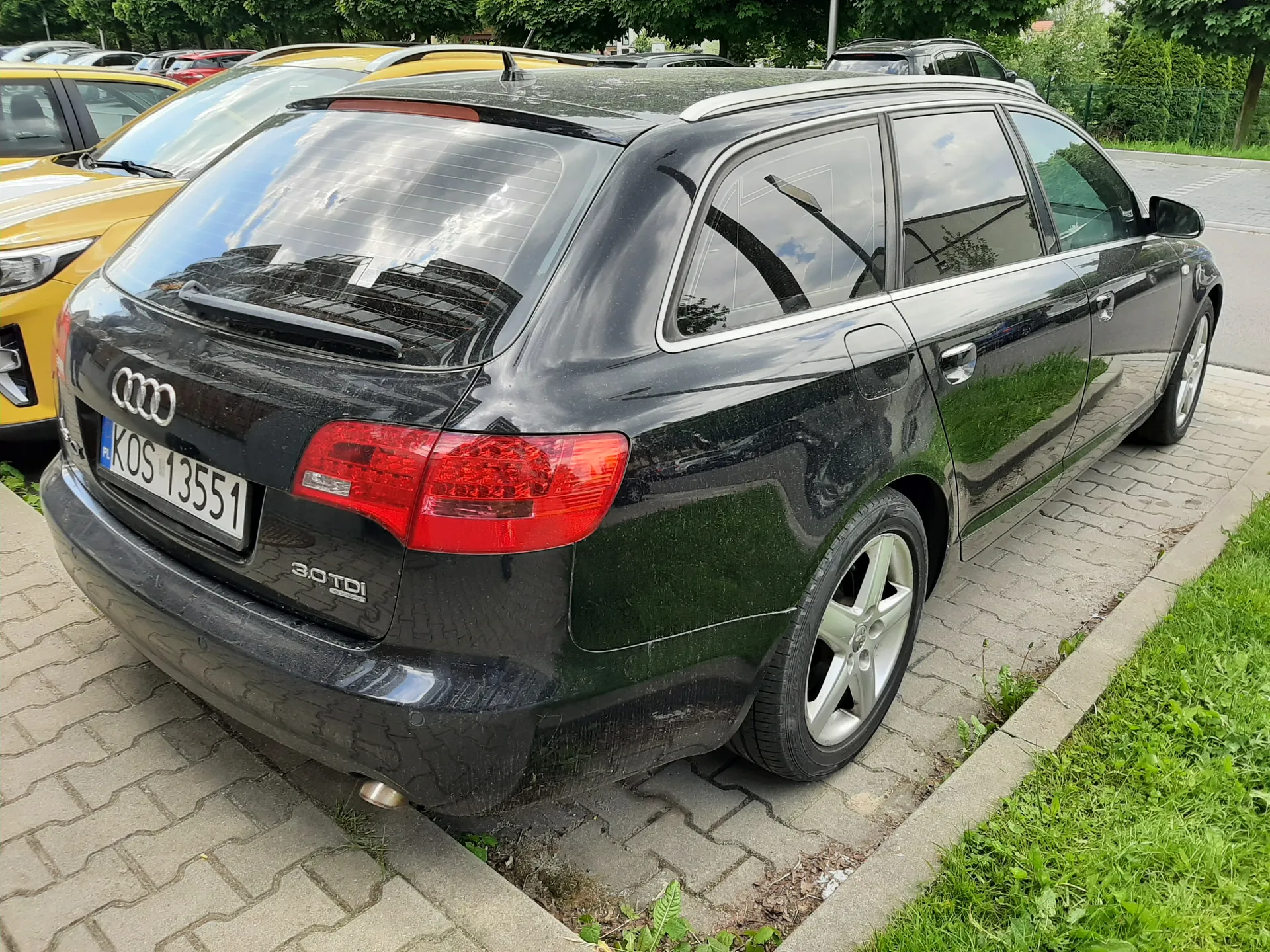 Czarny samochód Audi typu kombi widziany od tyłu z tablicą rejestracyjną KOS 13551 i napisem 3.0 TDI na bagażniku. Samochód zaparkowany na brukowanej powierzchni, obok żółtego auta. W tle widoczne inne pojazdy, drzewa oraz trawnik. Czerwone światła tylnego oświetlenia i srebrne felgi.
