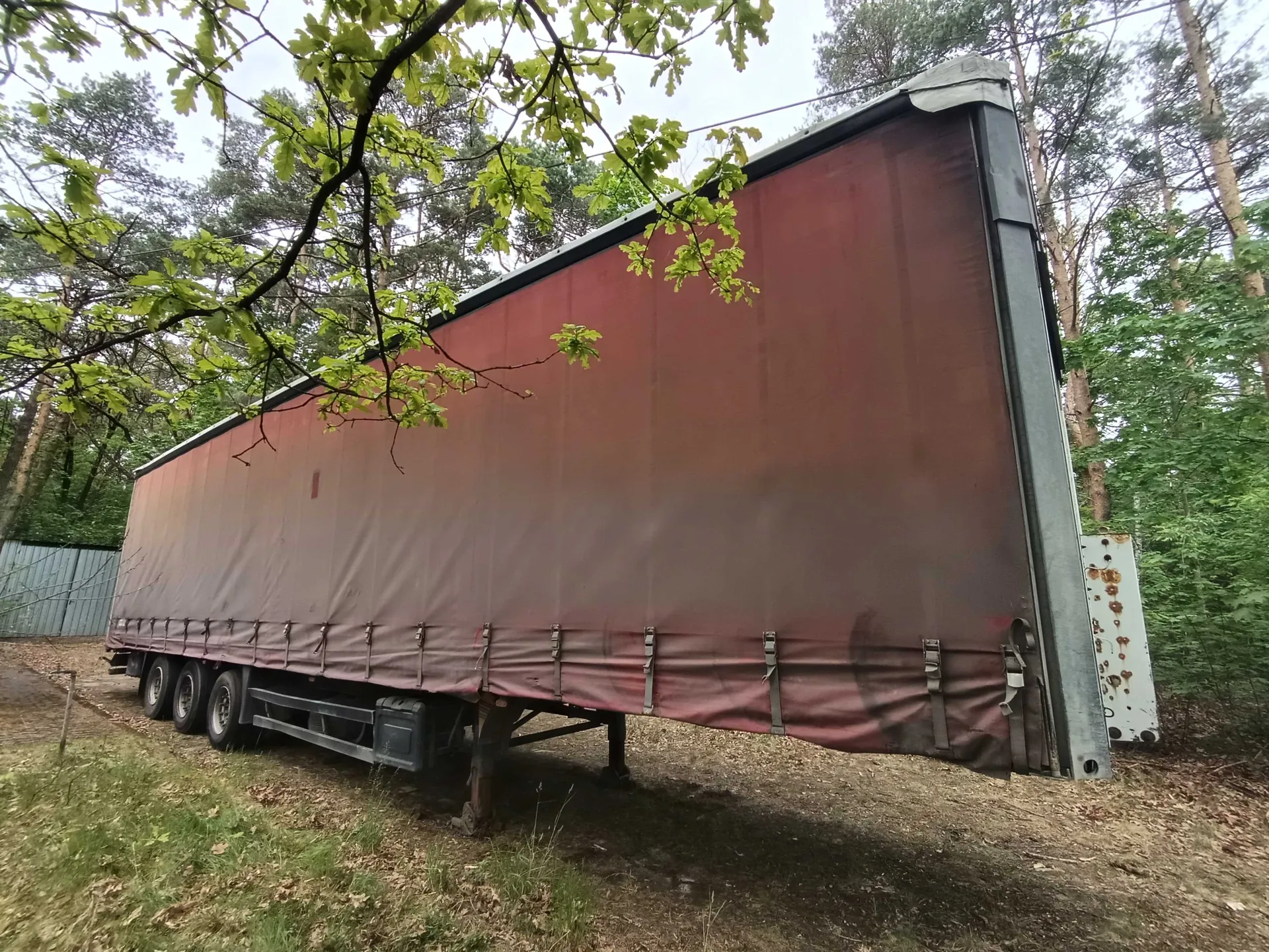 Duży przyczepa ciężarowa z brązowym namiotem, zaparkowana na leśnej polanie. Przyczepa ma metalowe elementy, zapięcia i koła. W tle widoczne są drzewa z zielonymi liśćmi, niebo oraz fragment metalowego ogrodzenia po lewej stronie. Podłoże składa się z trawy i suchych liści.