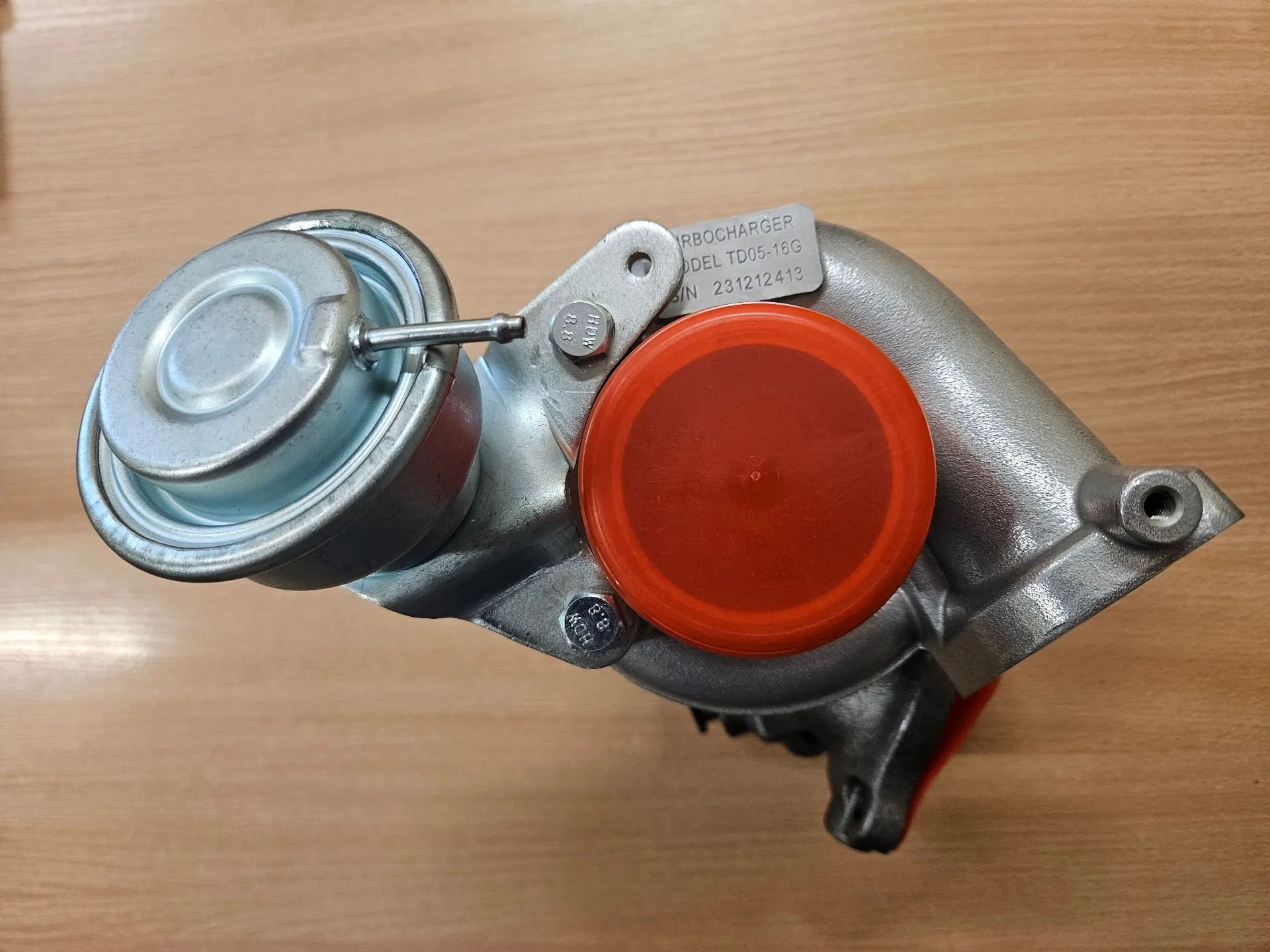 Metalowa turbosprężarka leżąca na drewnianej powierzchni. Urządzenie posiada srebrny korpus, okrągły element po lewej stronie oraz pomarańczową plastikową zaślepkę zamykającą jeden z wlotów. Na górnej części korpusu znajduje się srebrna tabliczka znamionowa z tekstem: 'TURBOCHARGER MODEL TD05-16G S/N 231212413'.