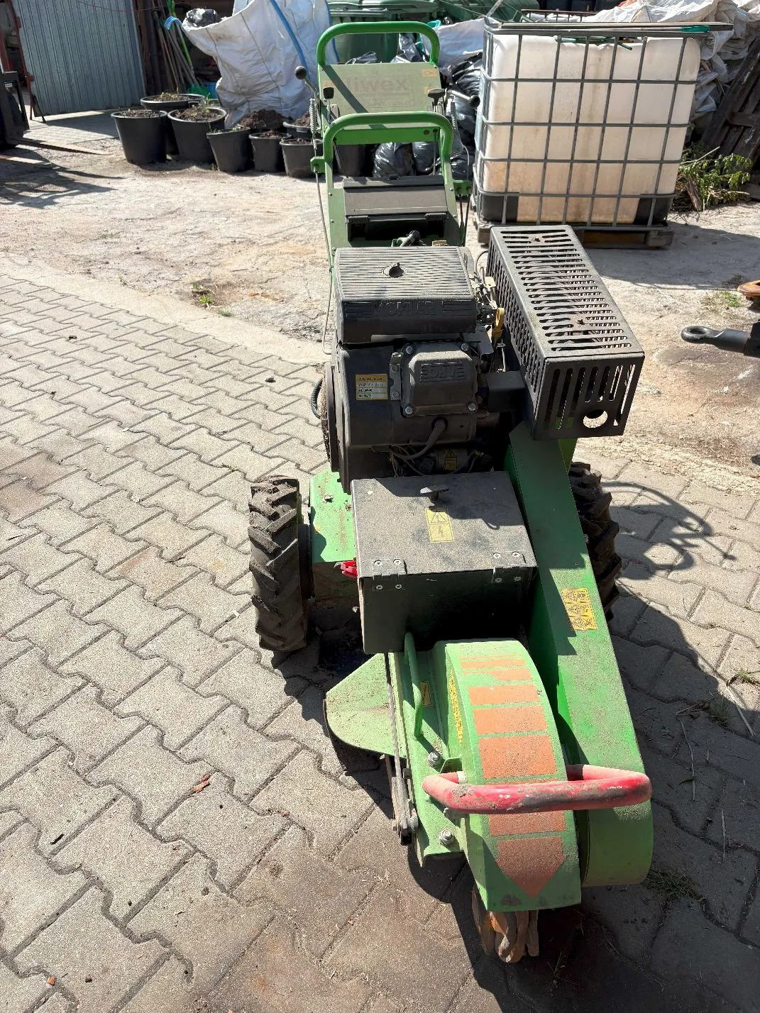 Zielona maszyna ogrodnicza, prawdopodobnie kosiarka do skarpy lub glebogryzarka, stojąca na kostce brukowej. Maszyna posiada silnik spalinowy, czarne opony z wyraźnym bieżnikiem oraz zieloną obudowę z czerwonym uchwytem z tyłu. W tle widoczne są czarne donice, białe worki oraz biały zbiornik IBC na palecie.