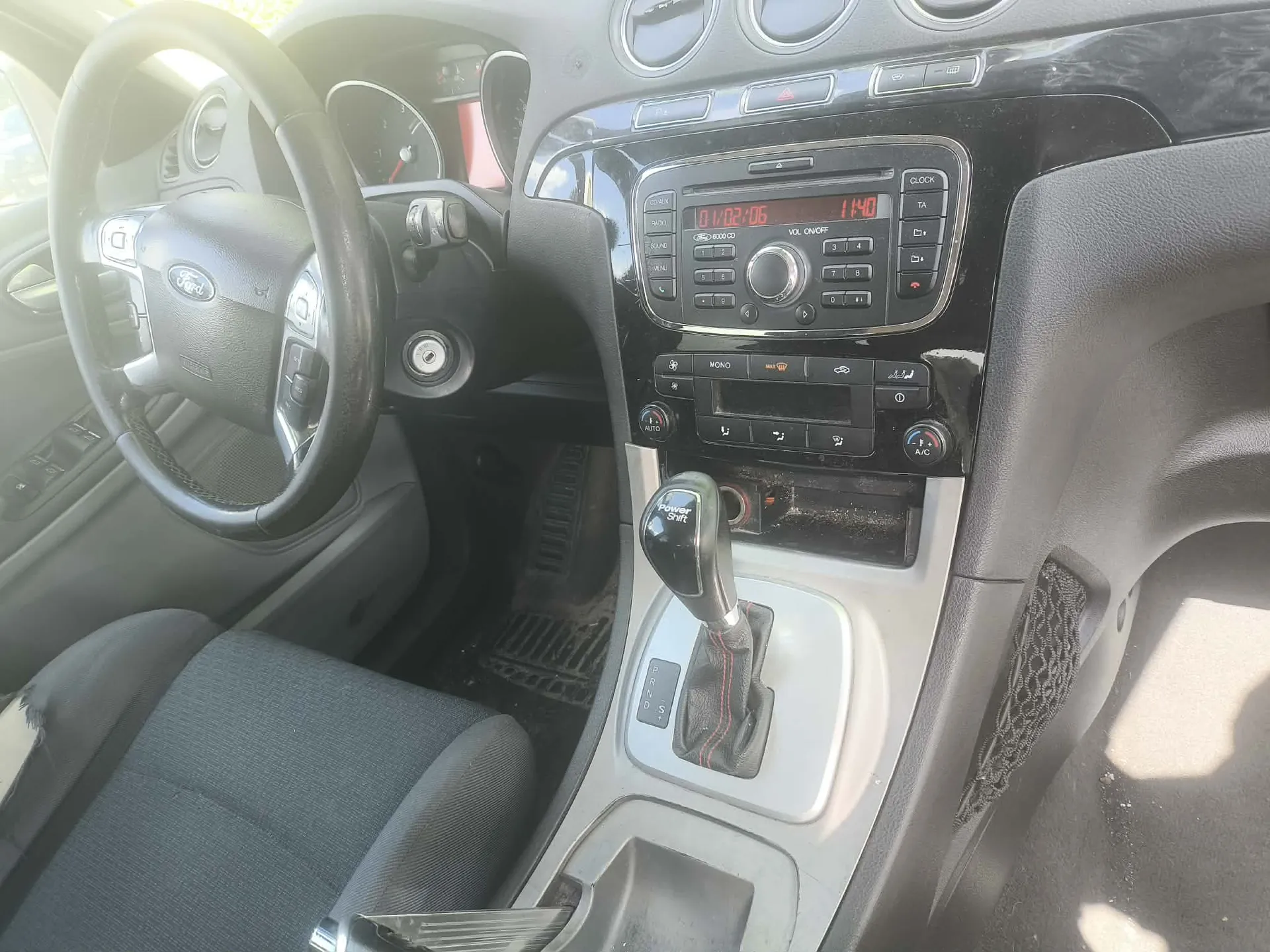 Wnętrze samochodu marki Ford, widok na deskę rozdzielczą i konsolę środkową. Po lewej stronie znajduje się kierownica z logo Forda. W centrum konsoli widoczne jest radio z czerwonym wyświetlaczem pokazującym godzinę 11:40 oraz datę 10/02/06. Poniżej radia znajdują się pokrętła i przyciski sterowania klimatyzacją. W dolnej części widoczna jest dźwignia zmiany biegów automatycznej skrzyni oraz fragment szarego materiałowego fotela kierowcy.