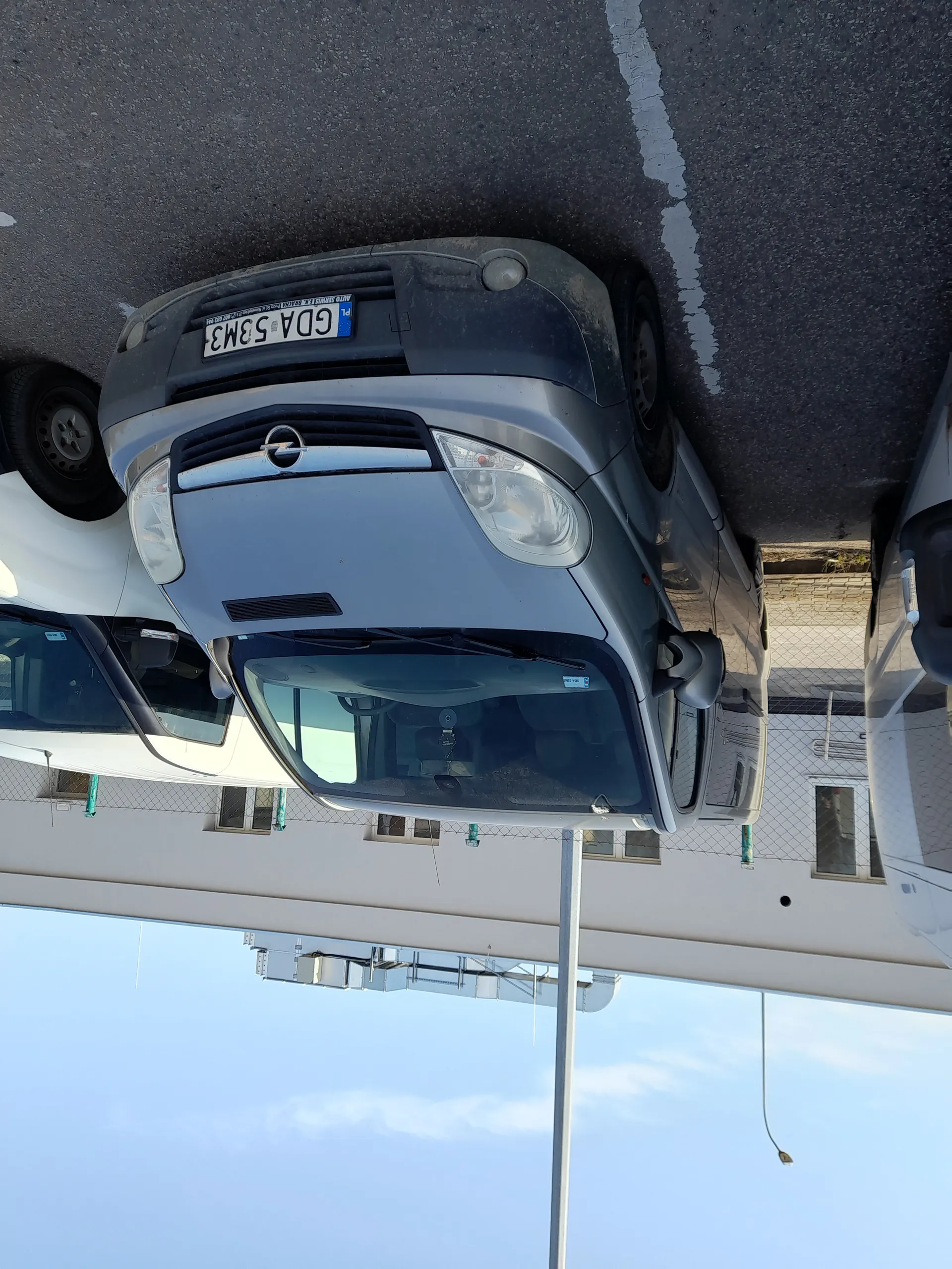 Dwa samochody zaparkowane na asfaltowym parkingu. Przedni szary samochód z tablicą rejestracyjną GDA 53M3. Za nim biały samochód. W tle budynek z oknami, ogrodzenie i latarnia uliczna. Niebo jest jasne z kilkoma chmurami.