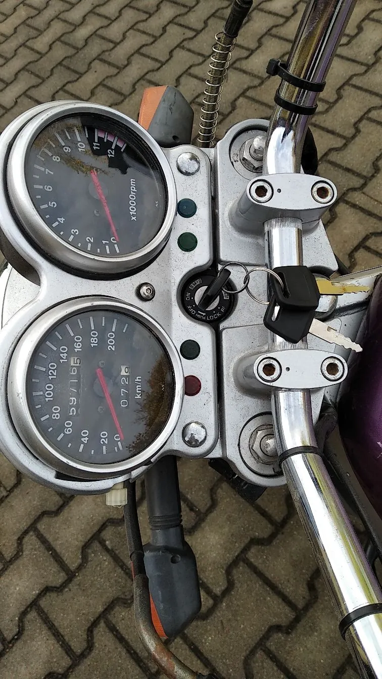 Widok z perspektywy kierowcy na kokpit motocykla. Widoczne są dwa okrągłe wskaźniki: górny to obrotomierz z oznaczeniem 'x1000rpm', a dolny to prędkościomierz z podziałką do 200 km/h. Pomiędzy wskaźnikami znajduje się stacyjka z włożonym kluczykiem i zawieszonym brelokiem. Po prawej stronie widoczna jest chromowana kierownica. Tło stanowi kostka brukowa.
