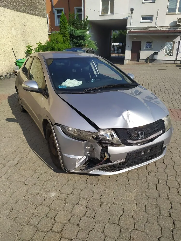 Srebrny samochód marki Honda z widocznymi uszkodzeniami przodu karoserii. Przedni zderzak jest częściowo oderwany i wgnieciony, a maska jest wygięta w górę. Pojazd stoi na kostce brukowej przed jasnym budynkiem mieszkalnym.