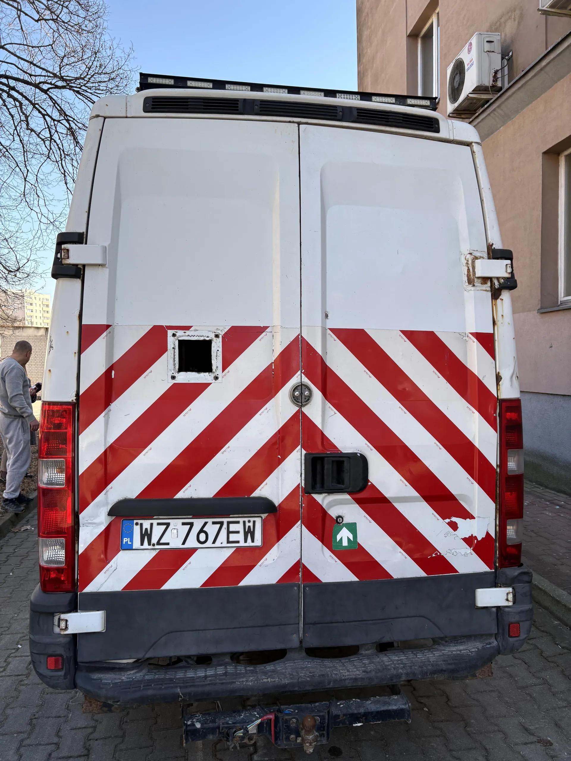 Tył białego furgonu z czerwonymi przekątnymi pasami ostrzegawczymi. Na środku tablica rejestracyjna z napisem WZ 767EW i niebieskim pasem z literą PL po lewej stronie. Po prawej stronie drzwi widoczny zielony znak z białą strzałką w górę. Furgon stoi na brukowanej ulicy obok budynku z klimatyzatorem. Po lewej stronie obrazu mężczyzna w szarej odzieży i bezlistne drzewo. Na tle błękitne niebo.