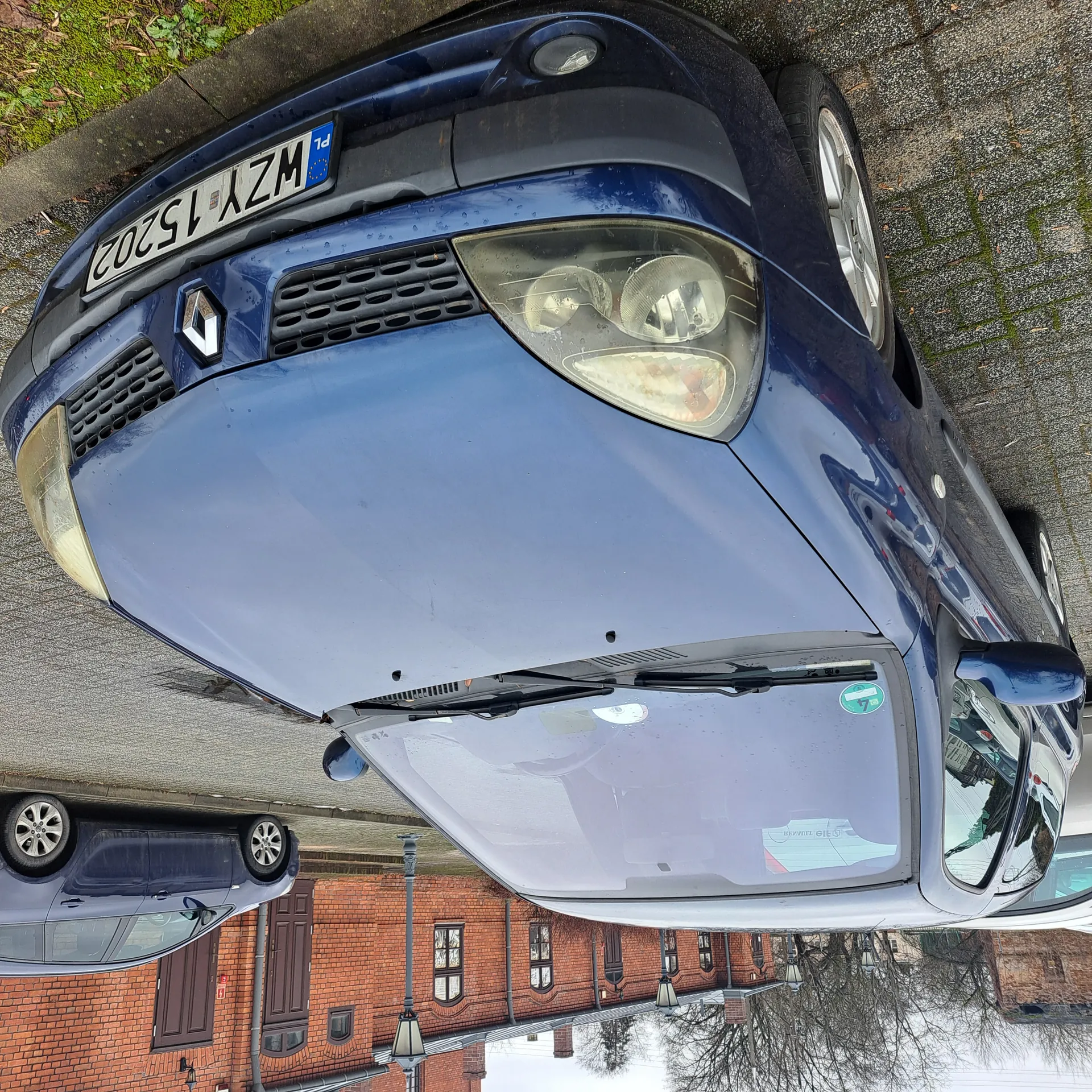 Niebieski samochód marki Renault z tablicą rejestracyjną WZY 15202 zaparkowany na żwirowanej powierzchni. W tle drugi ciemny samochód oraz ceglany budynek z brązowymi drzwiami i oknami. Na przedniej szybie widoczna zielona naklejka. Scena obejmuje brukowaną ulicę i drzewa bez liści.