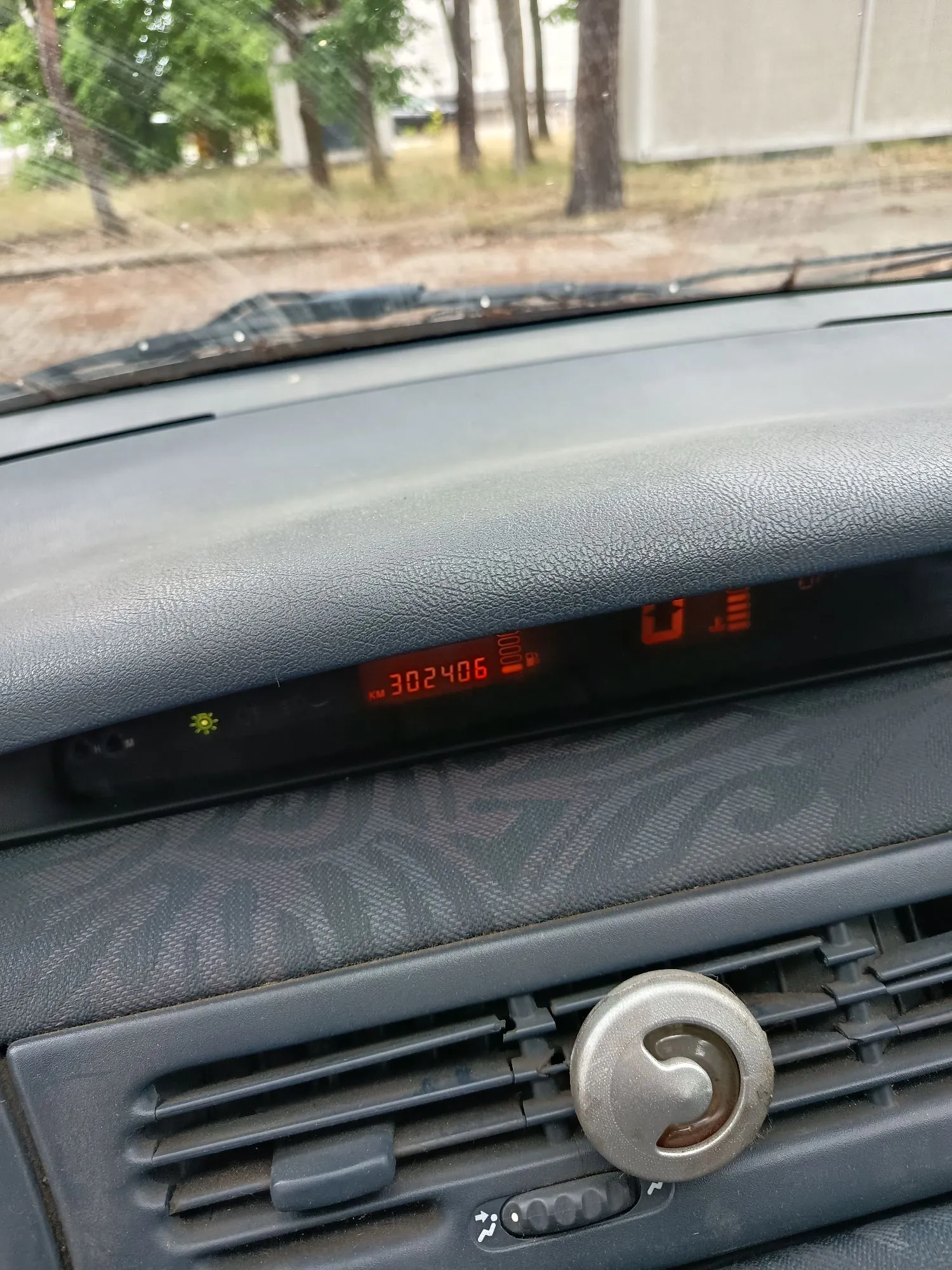 Widok z wnętrza samochodu na deskę rozdzielczą. W centralnej części znajduje się cyfrowy wyświetlacz z czerwonym tekstem wskazującym przebieg: 'KM 302406'. Obok wyświetlacza widoczne są kontrolki, w tym zapalona zielona ikona świateł. Poniżej znajduje się kratka nawiewu z okrągłym pokrętłem regulacyjnym.