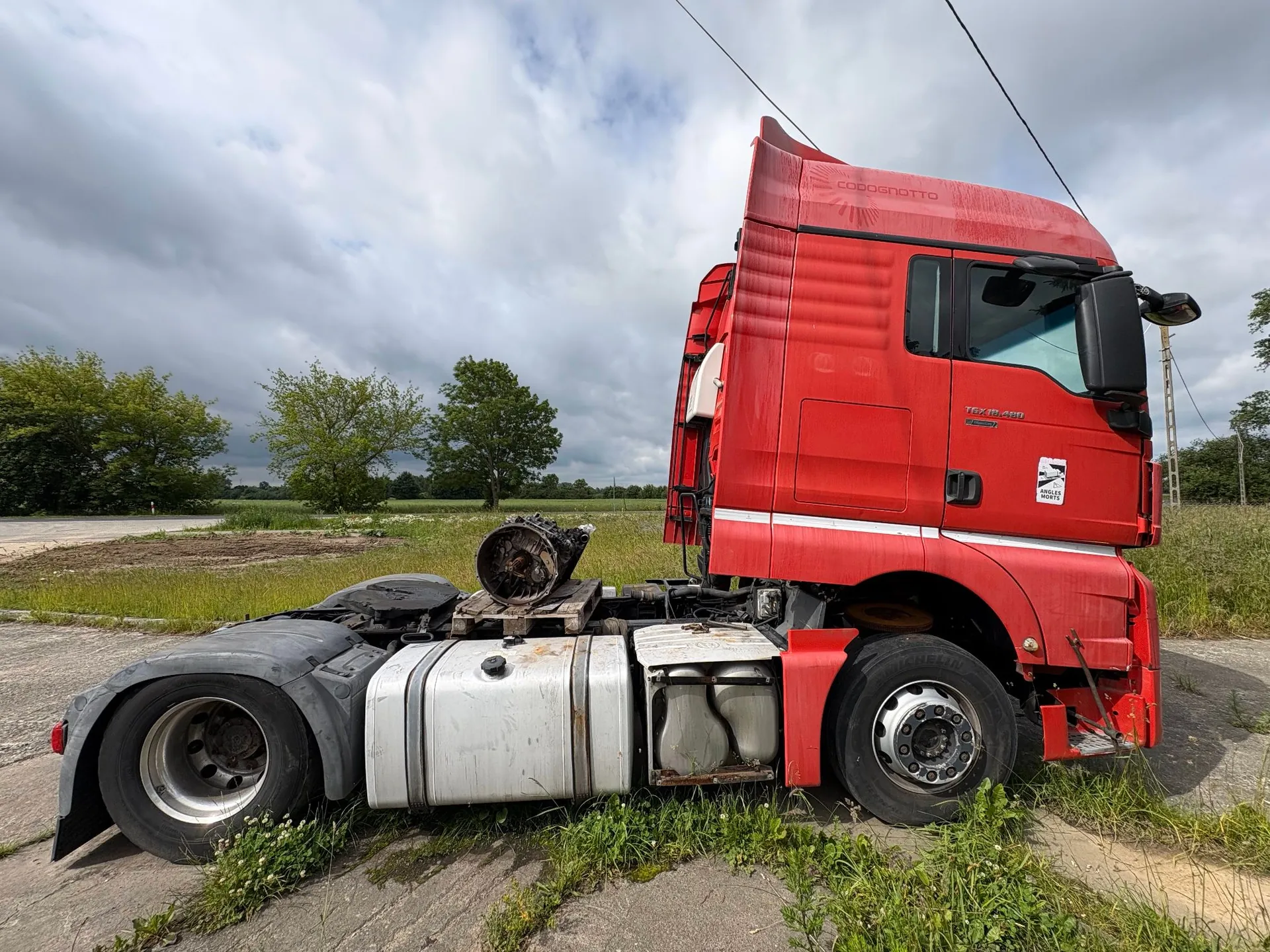Czerwona ciągnik siodłowy marki Scania, pozbawiony naczepy, zaparkowany na poboczu drogi. Na ramie pojazdu, w miejscu zazwyczaj zajmowanym przez naczepę, znajduje się metalowy element mechaniczny na drewnianej palecie. W tle widoczne są zielone drzewa, pola oraz zachmurzone niebo.