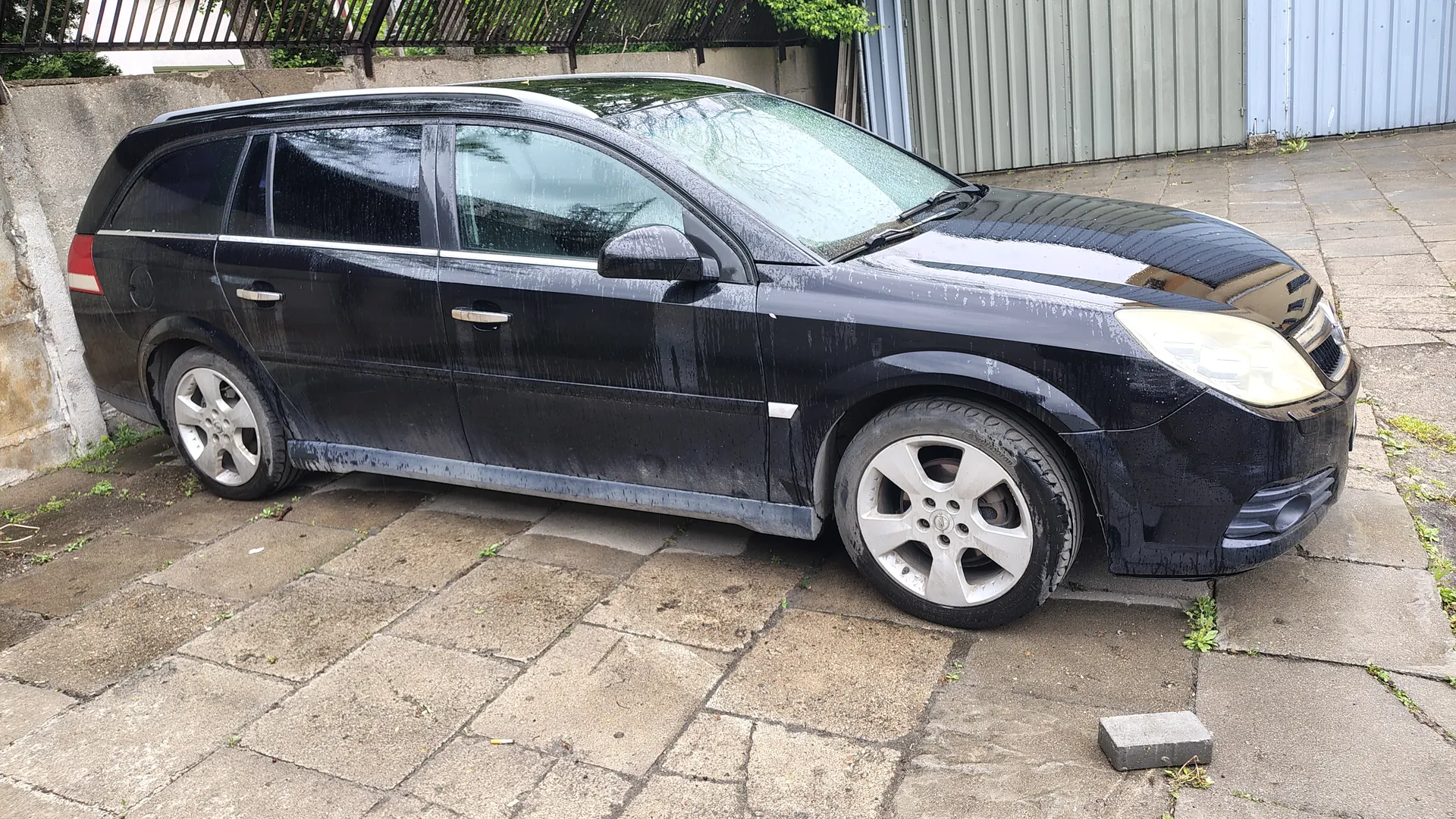 Czarny samochód typu kombi zaparkowany na brukowanej powierzchni. Pojazd pokryty jest kroplami wody, widoczne srebrne felgi i boczne lusterka. W tle betonowa ściana, metalowe ogrodzenie, zielone krzewy oraz metalowe budynki. Na ziemi leży mały betonowy blok.