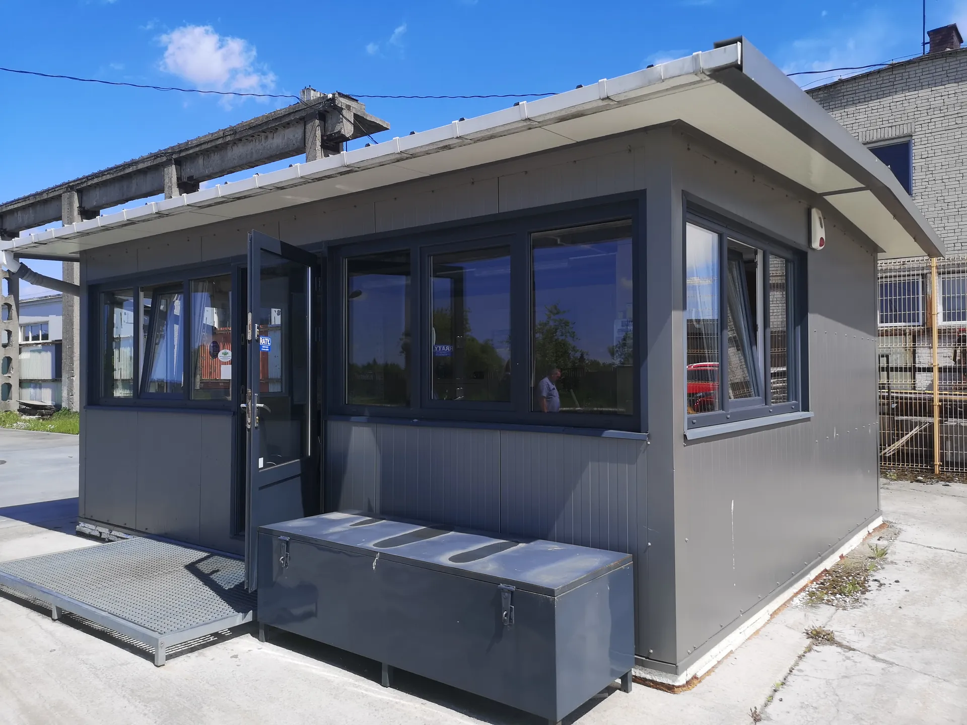 Szary, modułowy budynek strażniczy z ciemnymi ramami okiennymi i otwartymi drzwiami. Przed budynkiem znajduje się metalowa rampa oraz szary metalowy pojemnik. W tle widoczne są przemysłowe konstrukcje, budynek z cegły, siatka ogrodzeniowa oraz niebo z nielicznymi chmurami. Na drzwiach widoczny niebieski znak z napisem 'KATY'.