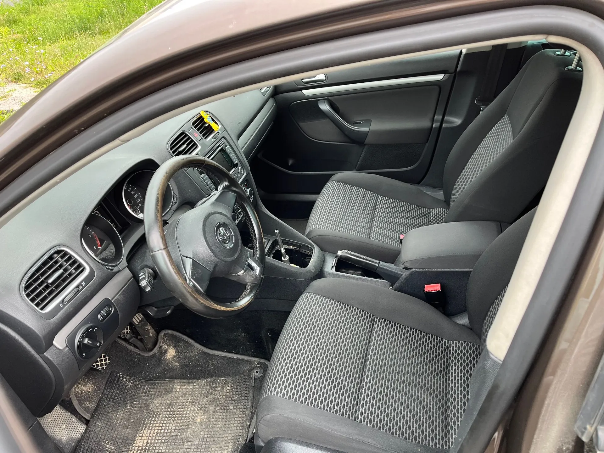 Wnętrze samochodu widziane z otwartych drzwi kierowcy. Widoczna jest czarna tapicerka materiałowa na fotelach, kierownica z logo Volkswagena, deska rozdzielcza z zegarami, system audio oraz dźwignia zmiany biegów. Na podłodze znajduje się gumowa mata z widocznymi zabrudzeniami z piasku.