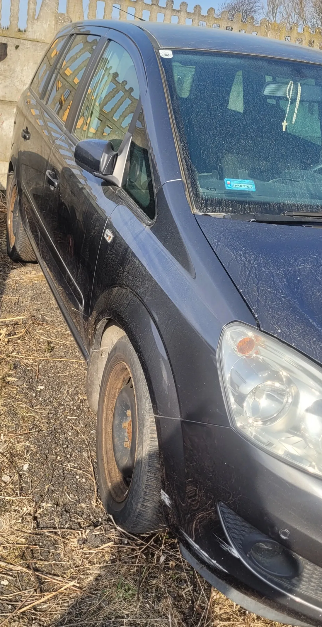 Czarny samochód osobowy zaparkowany na podłożu z żwirem i suchą trawą. Widoczne są zabrudzone koła, przedni reflektor oraz niebieski naklejony znaczek na przedniej szybie z napisem PKN 7757. Wewnątrz samochodu, na lusterku wstecznym, wiszą różaniec z krzyżem. W tle drewniane ogrodzenie i suche drzewa. Samochód ma ślady uszkodzeń na karoserii.