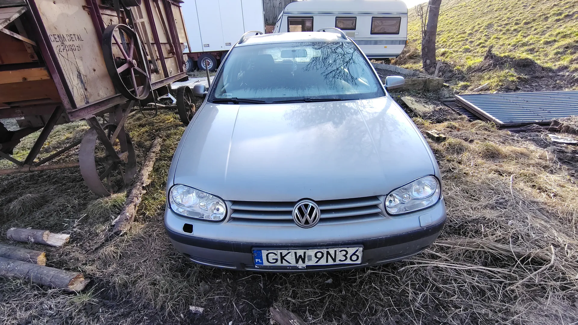 Srebrny samochód marki Volkswagen z tablicą rejestracyjną PL GKW 9N36, zaparkowany na trawiastym i błotnistym terenie. Po lewej stronie widoczny stary drewniany wóz z dużymi kołami i napisem CEMA SETAL 27000 21. W tle biały przyczepa lub naczepa z oknami oraz trawiasty stok. Na ziemi leżą kawałki drewna i metalowe elementy.