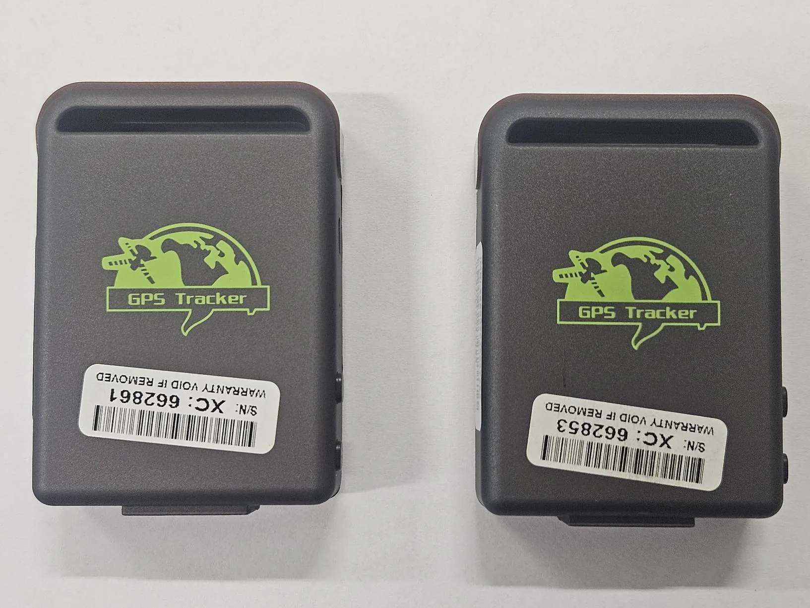 Dwa ciemnoszare urządzenia GPS Tracker ułożone obok siebie na białym tle. Każdy posiada zielony nadruk z napisem 'GPS Tracker' oraz ikoną globu z satelitą. Na każdym urządzeniu znajduje się biała etykieta z kodem kreskowym, numerem seryjnym (S/N: XC: 662861 i S/N: XC: 662853) oraz napisem 'WARRANTY VOID IF REMOVED', który jest umieszczony odwrócony do góry nogami. Urządzenia mają prostokątny kształt z wycięciem na górze.