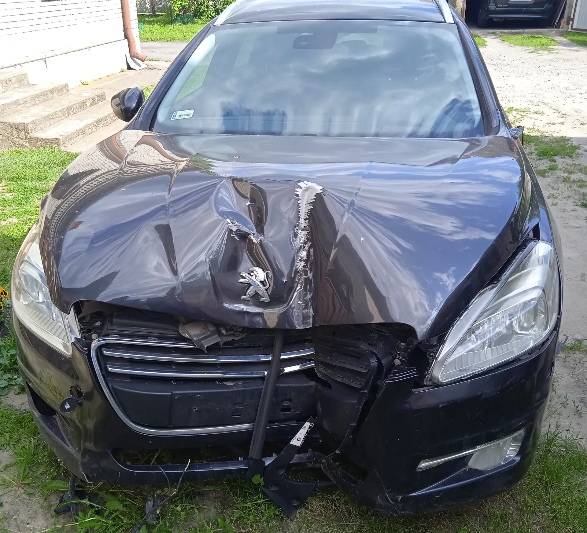 Czarny samochód marki Peugeot z poważnie uszkodzonym przodem. Widoczne zgniecione maski, uszkodzona atrapa chłodnicy, pęknięte reflektory oraz częściowo odsłonięte elementy pod maską. Na masce znajduje się srebrny emblemat marki. Samochód stoi na trawie, w tle budynek z białymi schodami i częściowo widoczny inny pojazd. Na przedniej szybie widoczny jest mały naklejony dokument.