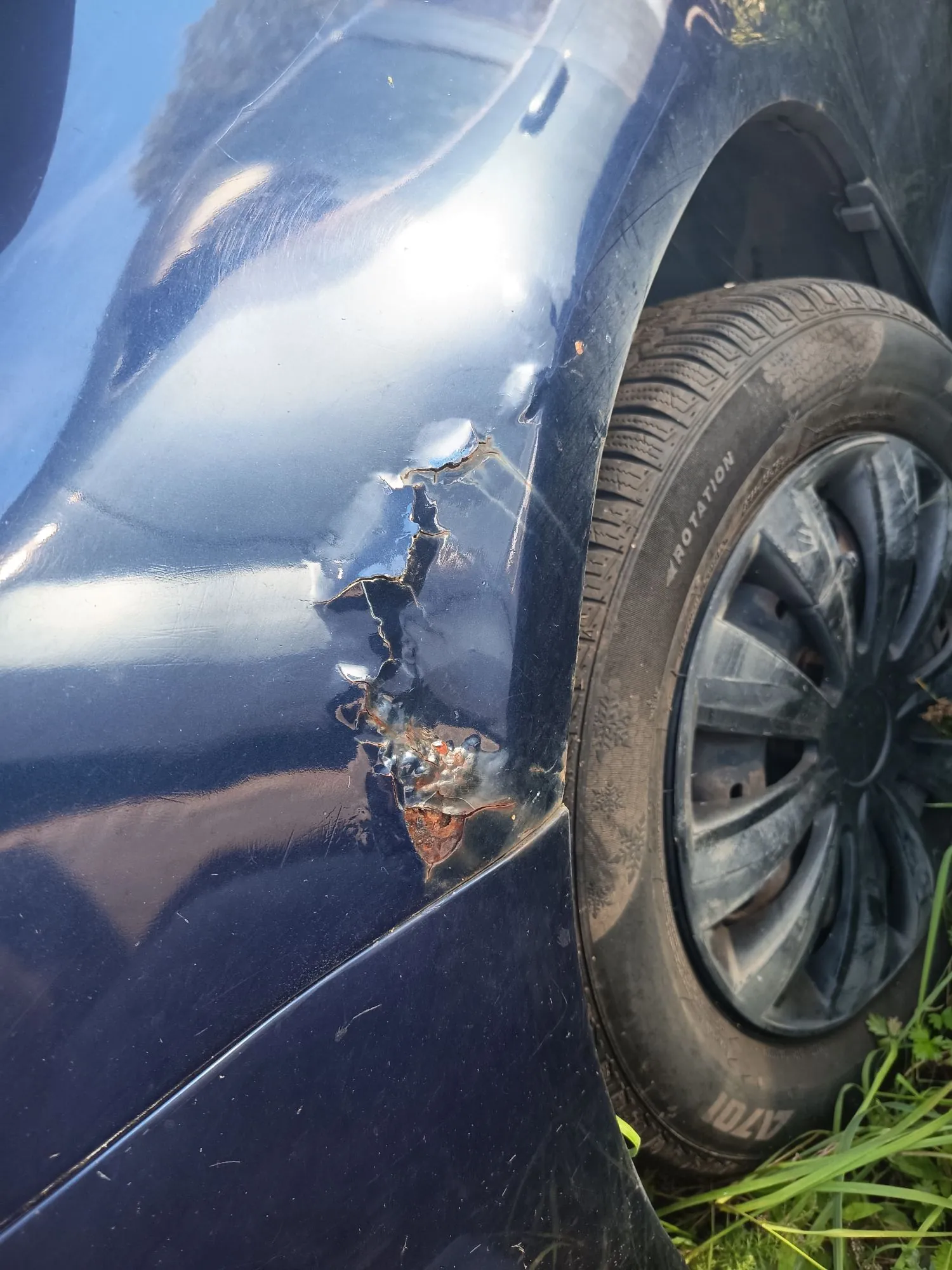 Zbliżenie na uszkodzony element nadwozia ciemnoniebieskiego samochodu w okolicy nadkola. Blacha jest wgnieciona, z odpryskami lakieru i widocznymi śladami korozji (rdzy) w miejscu uszkodzenia. Obok znajduje się czarna opona z felgą.