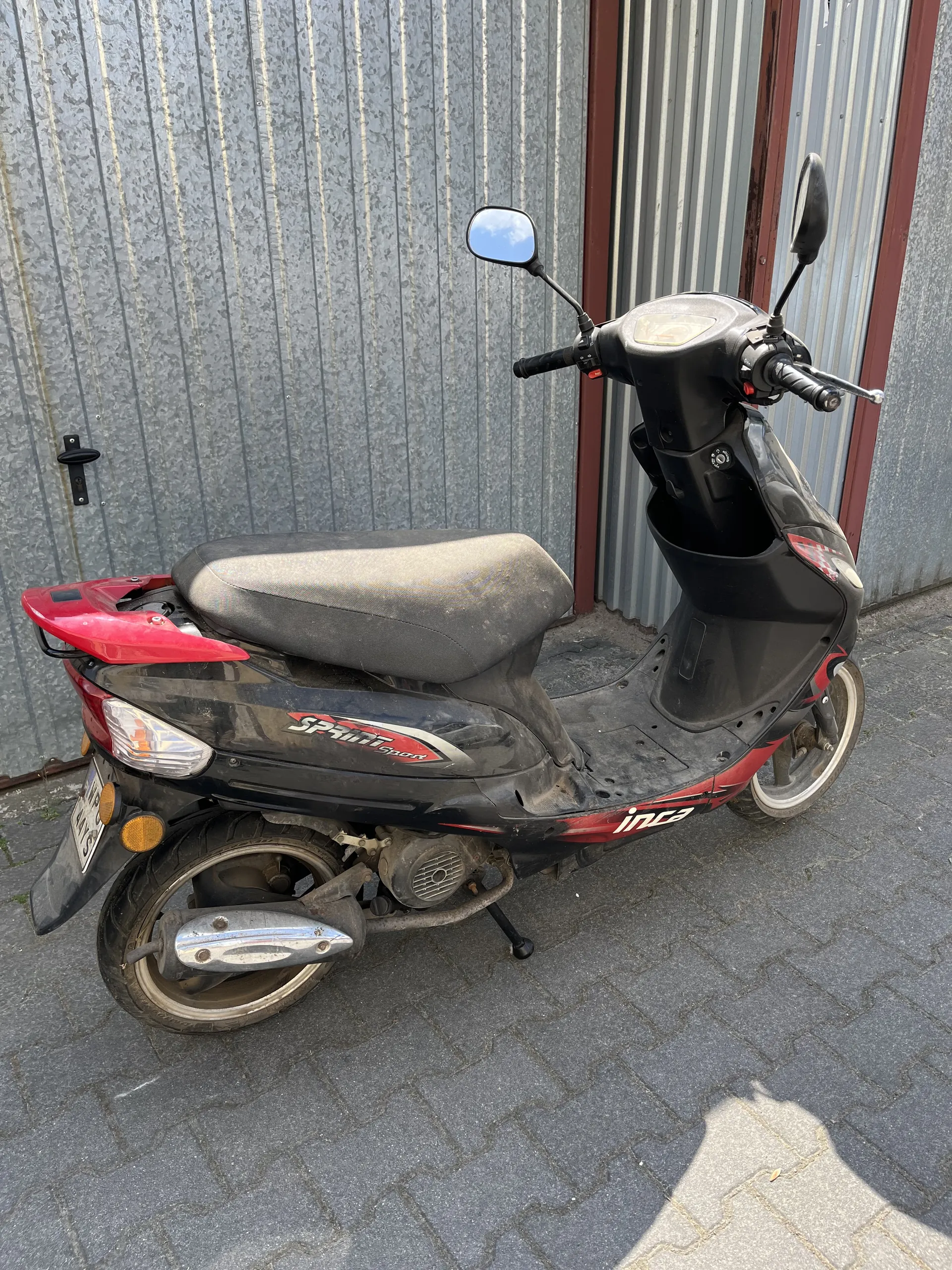 Czarno-czerwony skuter zaparkowany na brukowanej powierzchni obok budynku z metalową bramą garażową. Na skuterze widoczne napisy 'Spirit' oraz 'ing'. Skuter posiada czarne siedzenie, lusterka boczne i tablicę rejestracyjną. Brama garażowa ma czerwone wykończenie.