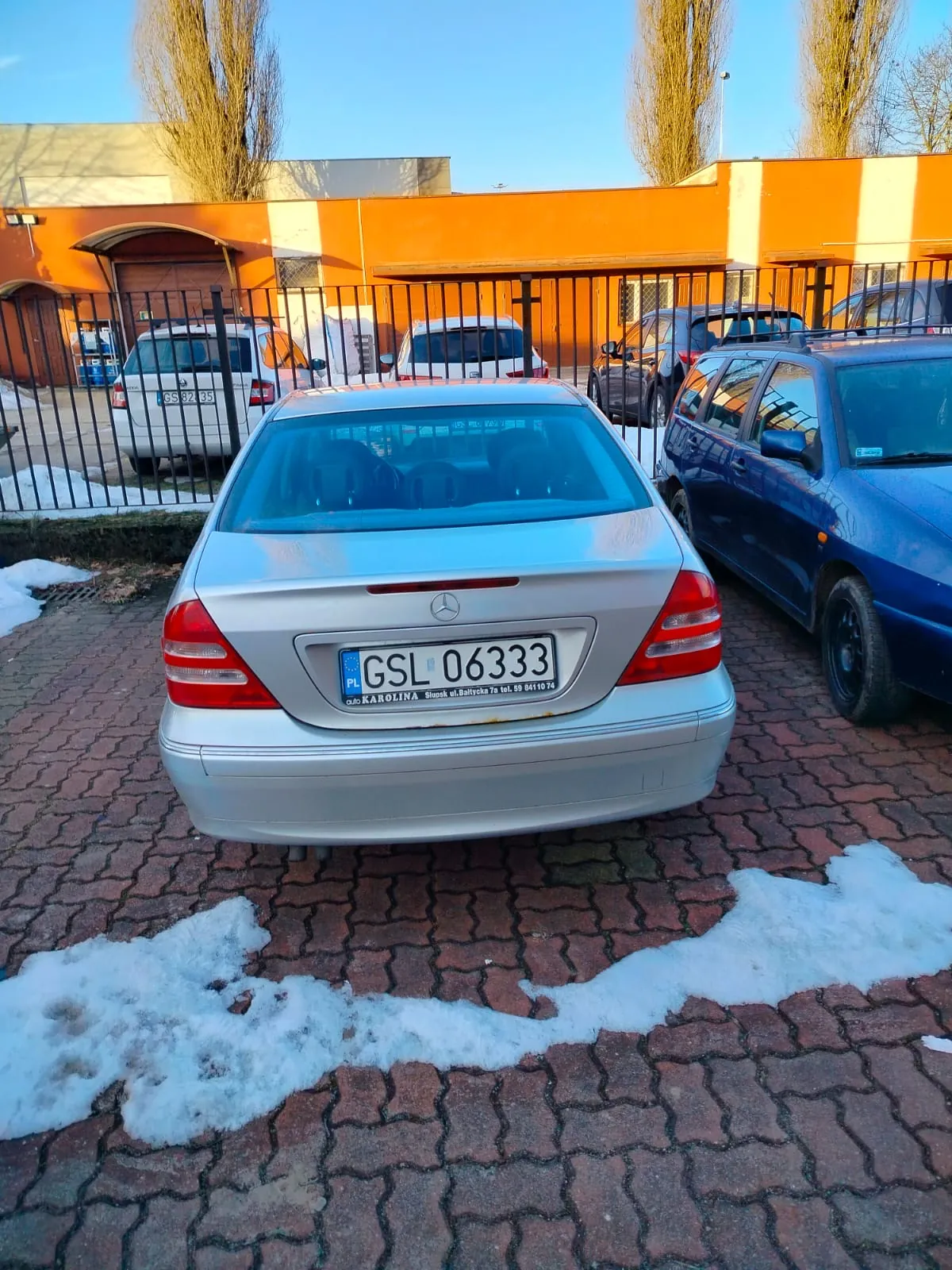 Zdjęcie przedstawia srebrny samochód Mercedes-Benz z tyłu, zaparkowany na brukowanej powierzchni. Na tablicy rejestracyjnej widoczny jest napis "PL GSL 06333" oraz dodatkowy tekst "KAROLINA" i adres. W tle znajdują się inne pojazdy: biały van, niebieski samochód osobowy oraz czarny samochód. Za ogrodzeniem metalowym widoczne są pomarańczowe budynki, drzewa bez liści i resztki śniegu na ziemi. Niebo jest jasne, a na brukowanych placach znajdują się plamy śniegu.