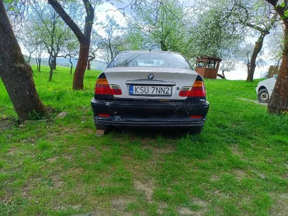Tył srebrnego samochodu marki BMW z tablicą rejestracyjną KSU 7NN2, zaparkowanego na trawiastym obszarze z widocznymi plamami ziemi. W tle drzewa z zielonymi liśćmi, częściowo widoczny biały samochód po prawej stronie oraz drewniana konstrukcja. Niebo jest częściowo zachmurzone.