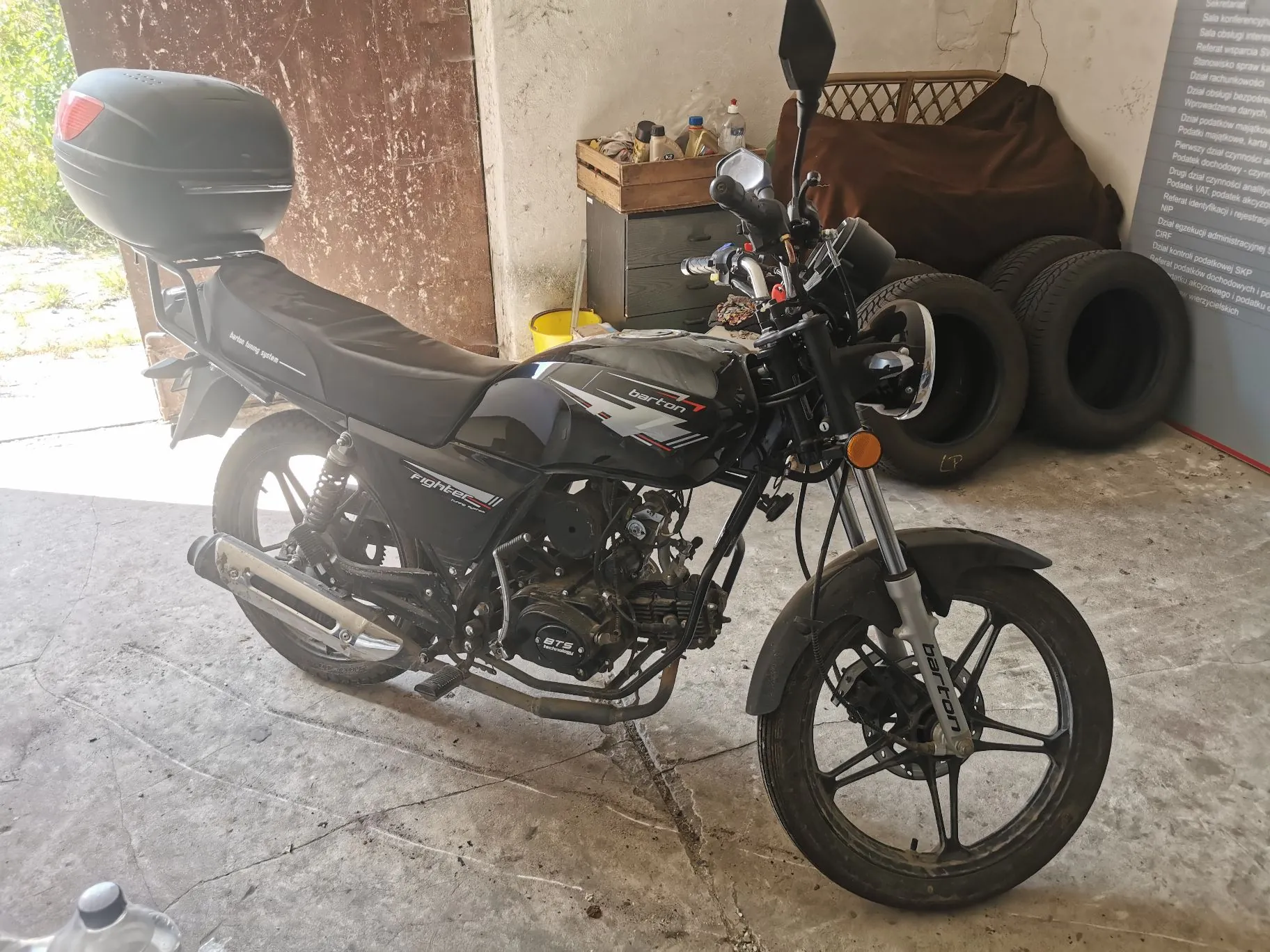 Czarny motocykl marki Barton model Fighter, zaparkowany w garażu na betonowej podłodze. Motocykl posiada czarne felgi, czarną kanapę oraz zamontowany z tyłu czarny kufer. Na baku widnieje napis 'barton', a na bocznej owiewce napis 'Fighter'. W tle widoczne są opony samochodowe, szafka narzędziowa oraz fragmenty białej ściany z tekstem.