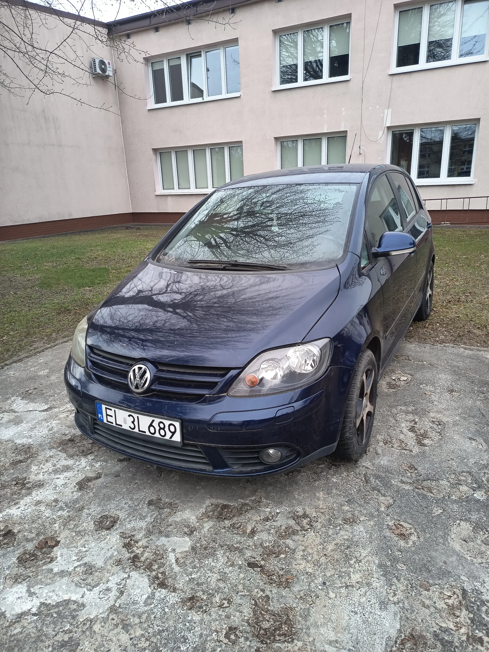 Niebieski samochód marki Volkswagen z tablicą rejestracyjną EL 3L689 (PL) zaparkowany na betonowym placu przed jasnoróżowym budynkiem z białymi oknami. W tle widoczne są suche gałęzie drzew i zielona trawa.