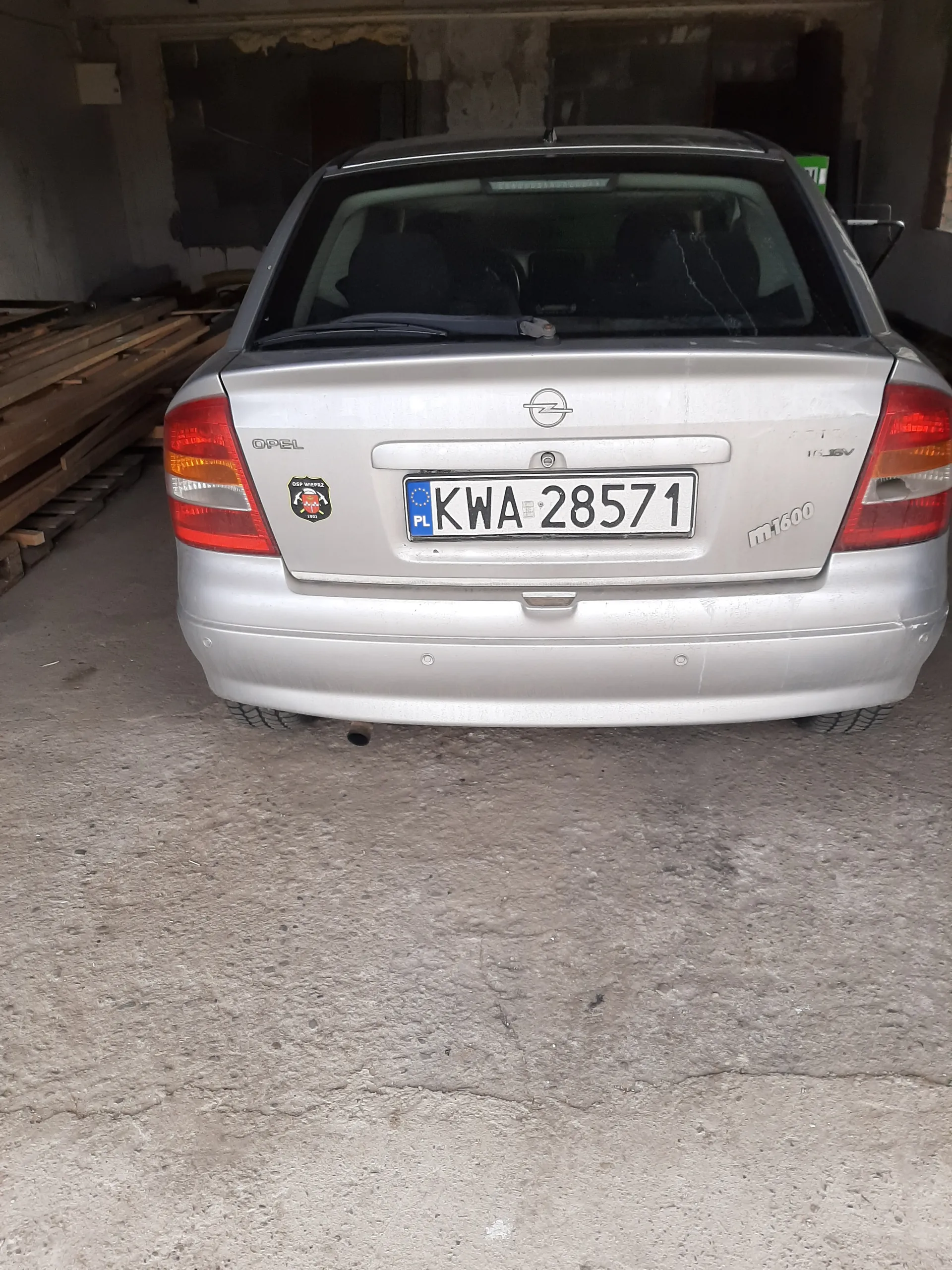 Srebrny samochód Opel z tyłu, z tablicą rejestracyjną KWA 28571 z niebieskim pasem PL po lewej stronie. Na bagażniku widoczne logo Opel, napis 1600 oraz naklejka. Samochód zaparkowany na betonowej powierzchni, w tle budynek z drewnianymi deskami.