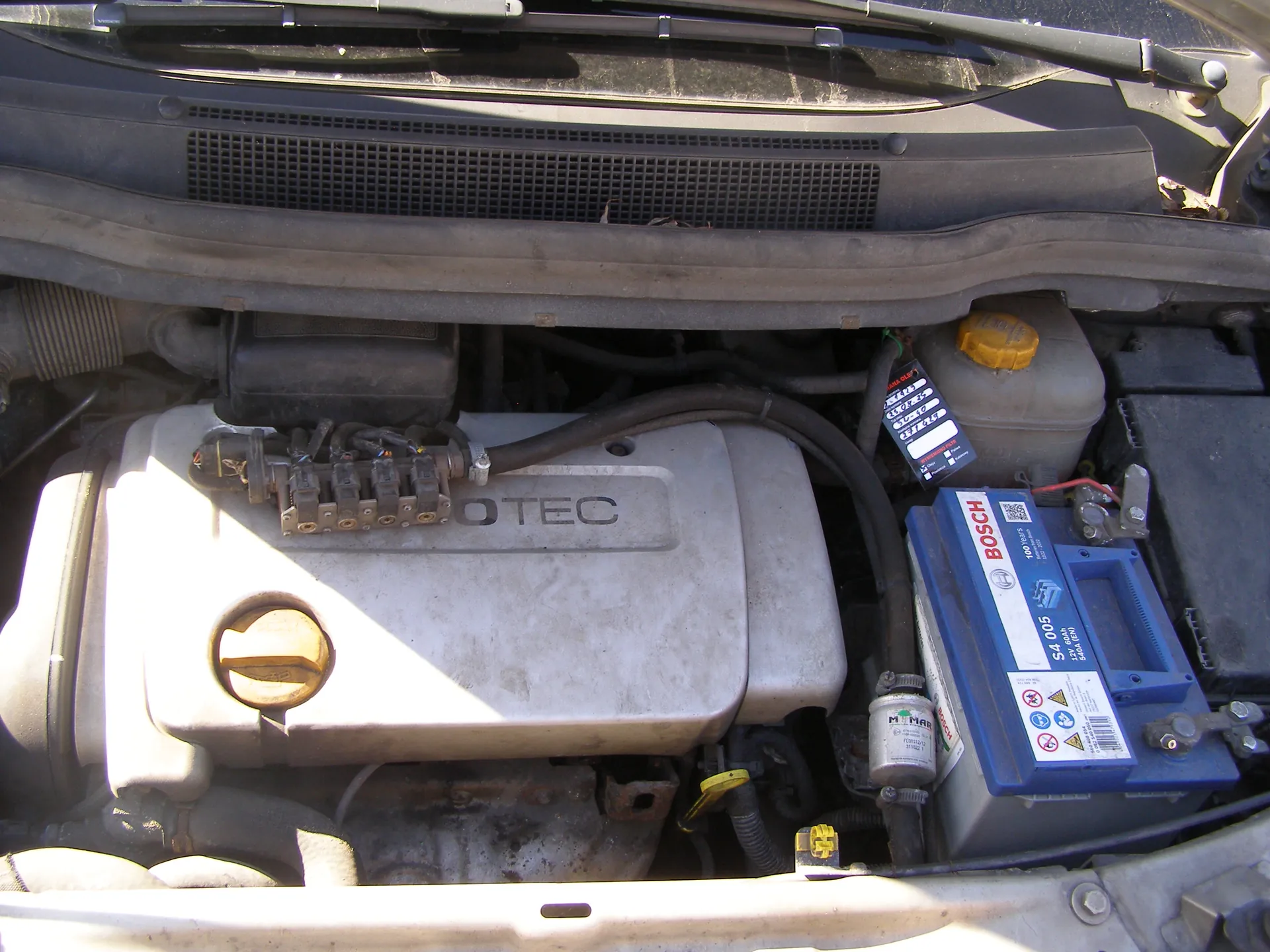 Widok komory silnika samochodu z otwartą maską. Centralnie umieszczona pokrywa silnika z napisem 'ECOTEC'. Po prawej stronie bateria marki BOSCH z oznaczeniem 'S4 005' oraz żółtymi nakrętkami. Widoczne są również inne elementy, takie jak przewody, zbiornik z żółtą pokrywką oraz siatka wentylacyjna pod przednią szybą.