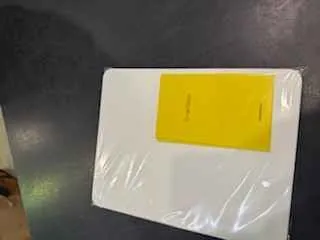 Biały prostokątny przedmiot owinięty przezroczystą folią, z żółtym prostokątnym etykietą na górze. Tło stanowi ciemna powierzchnia. Na etykiecie widoczny jest nieczytelny tekst.