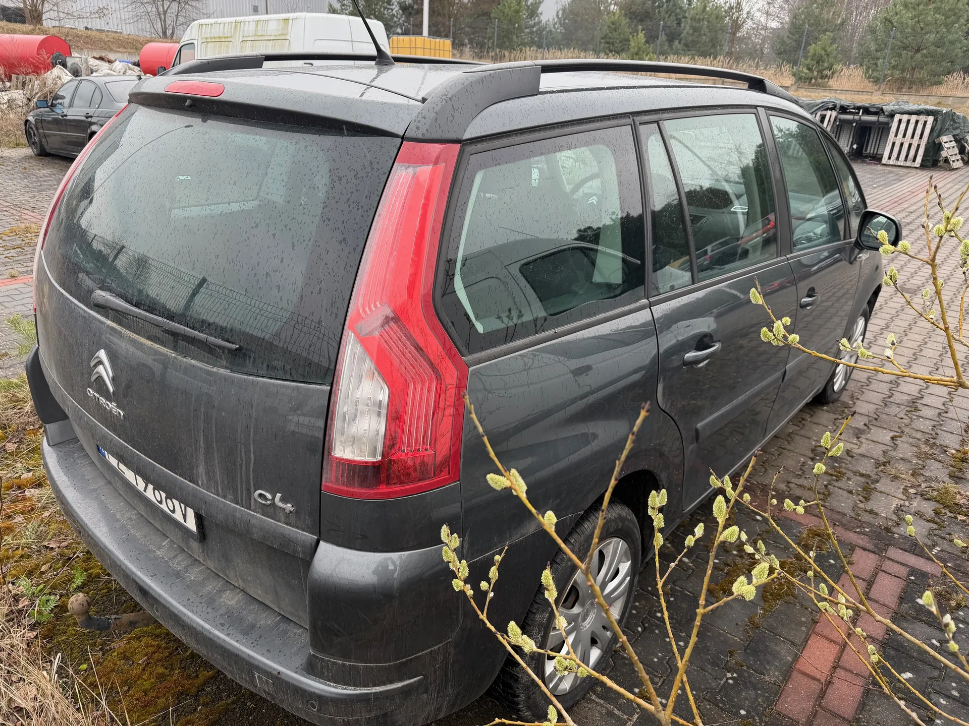 Ciemnoszary samochód Citroën C4 Picasso zaparkowany na brukowanej powierzchni. Widoczny tył pojazdu z czerwonymi światłami, logo Citroën, napisem C4 i tablicą rejestracyjną EL 1766V. Na szybie tylnej krople deszczu. W tle inne pojazdy (ciemny samochód, biały furgon), czerwone zbiorniki, stosy materiałów i drzewa. W pierwszym planie gałęzie z zielonymi pąkami. Samochód ma dachowe pręty i ciemne szyby.