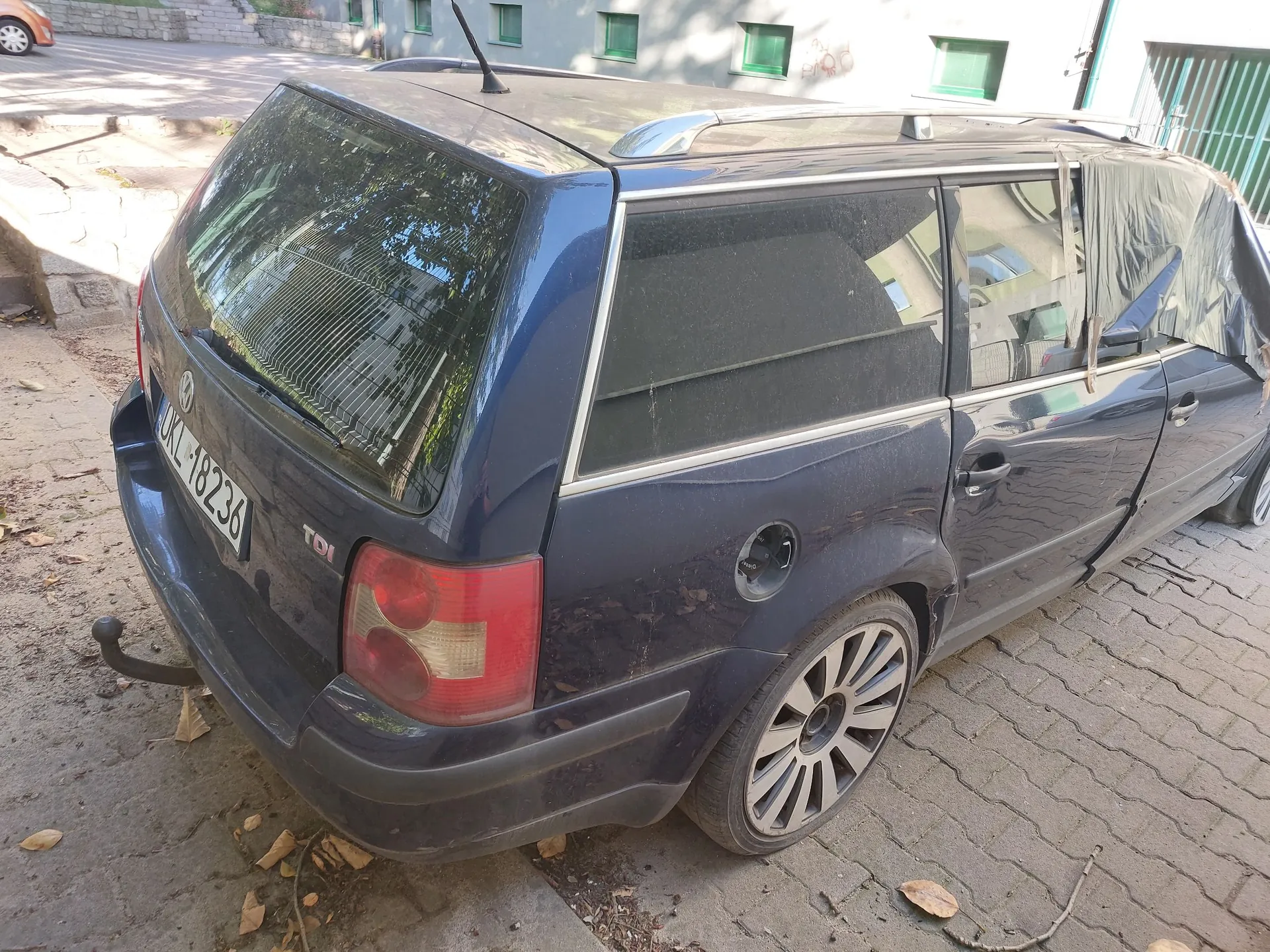 Tylna i boczna część granatowego samochodu marki Volkswagen w nadwoziu kombi, zaparkowanego na kostce brukowej. Pojazd jest zakurzony, posiada hak holowniczy oraz tablicę rejestracyjną o numerze UKL 18236. Na klapie bagażnika widnieje oznaczenie TDI. W tle widać fragment jasnego budynku z zielonymi ramami okiennymi.