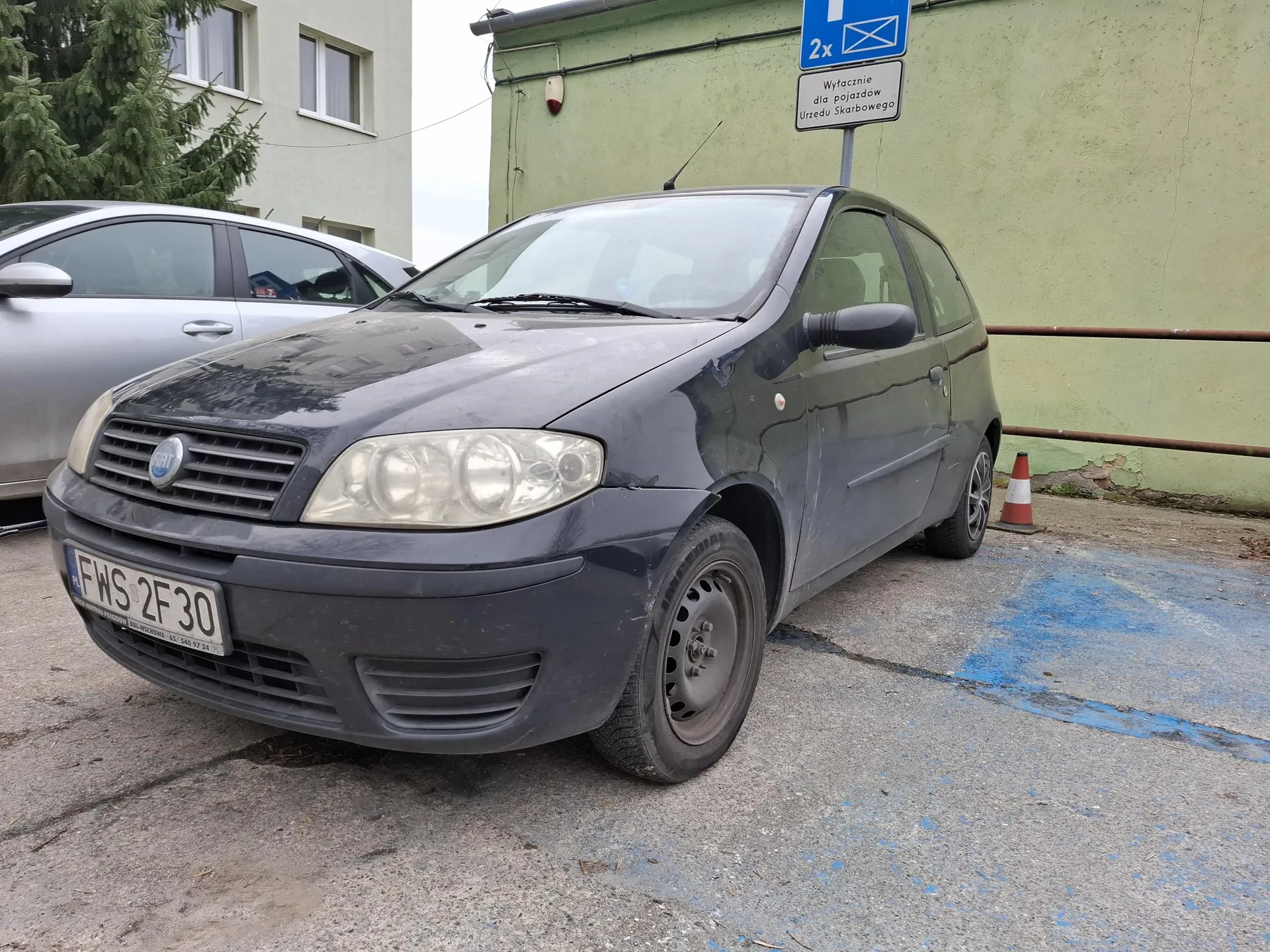 Ciemnoszary samochód marki Fiat z tablicą rejestracyjną FWS 2F30 zaparkowany na betonowym placu. W tle zielony budynek, srebrny samochód, drzewo oraz niebieski znak parkingowy z napisem '2x' i tabliczką 'Wyłącznie dla pojazdów Urzędu Skarbowego'. Obok samochodu stoi pomarańczowo-biały stożek drogowy, a na ziemi widoczne są niebieskie linie malowane na asfalcie.