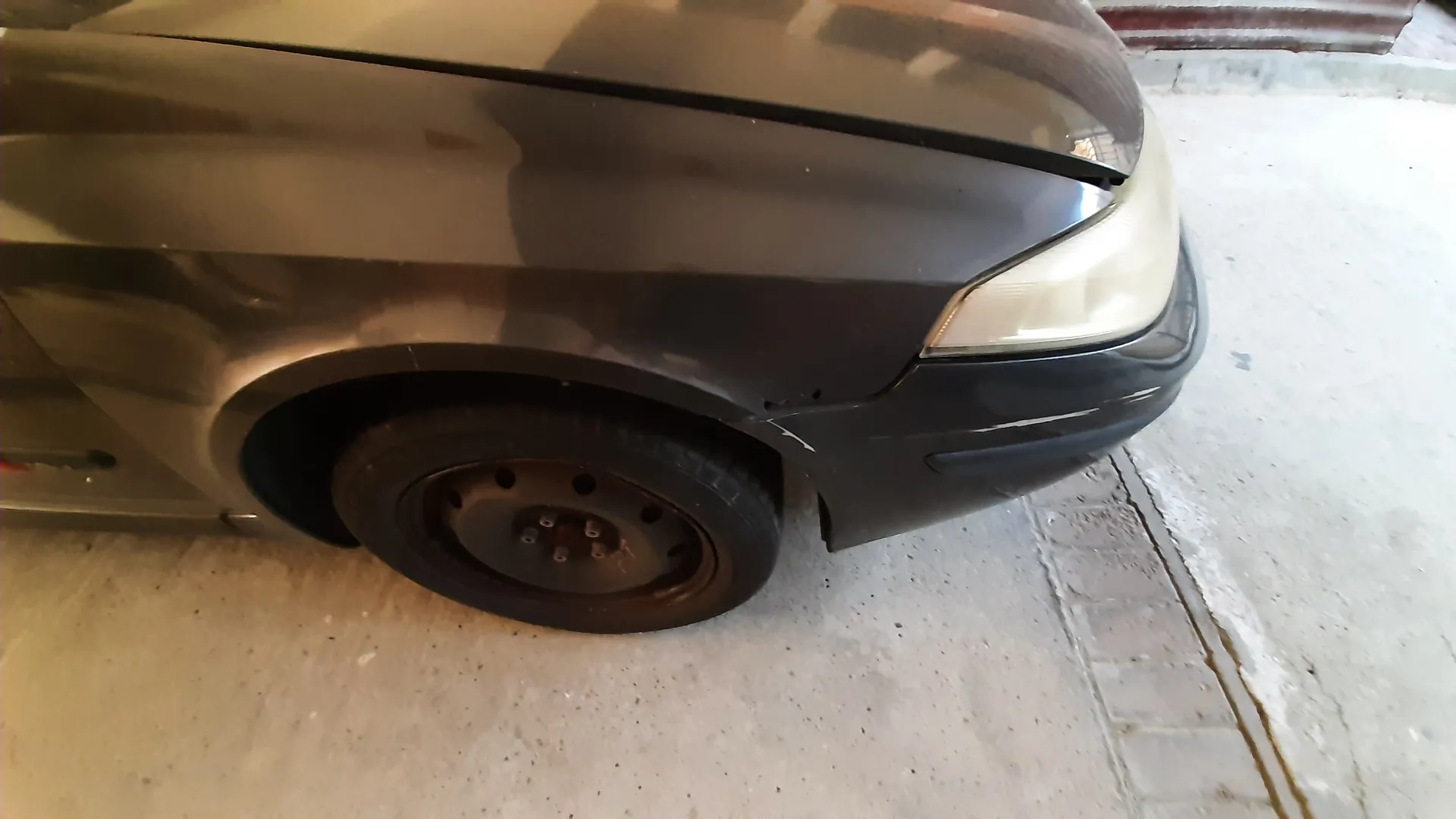 Przednia prawa część ciemnoszarego samochodu z widocznym reflektorem, kołem z brązowym felgą stalową i oponą. Samochód stoi na betonowej powierzchni z ceglanym wykończeniem po prawej stronie. W tle widoczny fragment chodnika i krawężnika.