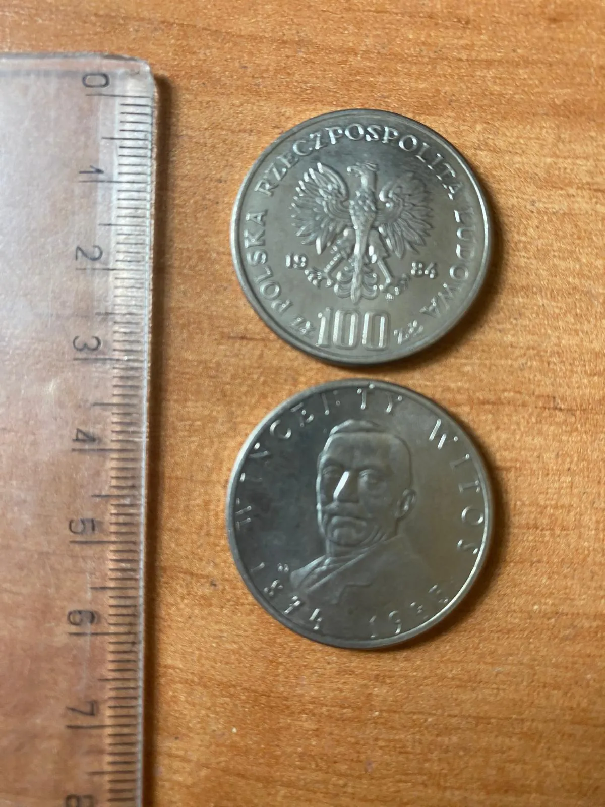 Dwie srebrzyste monety leżące obok metalowej linijki na drewnianym blacie. Górna moneta przedstawia orła w koronie z napisem 'RZECZPOSPOLITA LUDOWA POLSKA' oraz nominałem '100' i datą '1984'. Dolna moneta przedstawia profil mężczyzny w średnim wieku z wąsami.