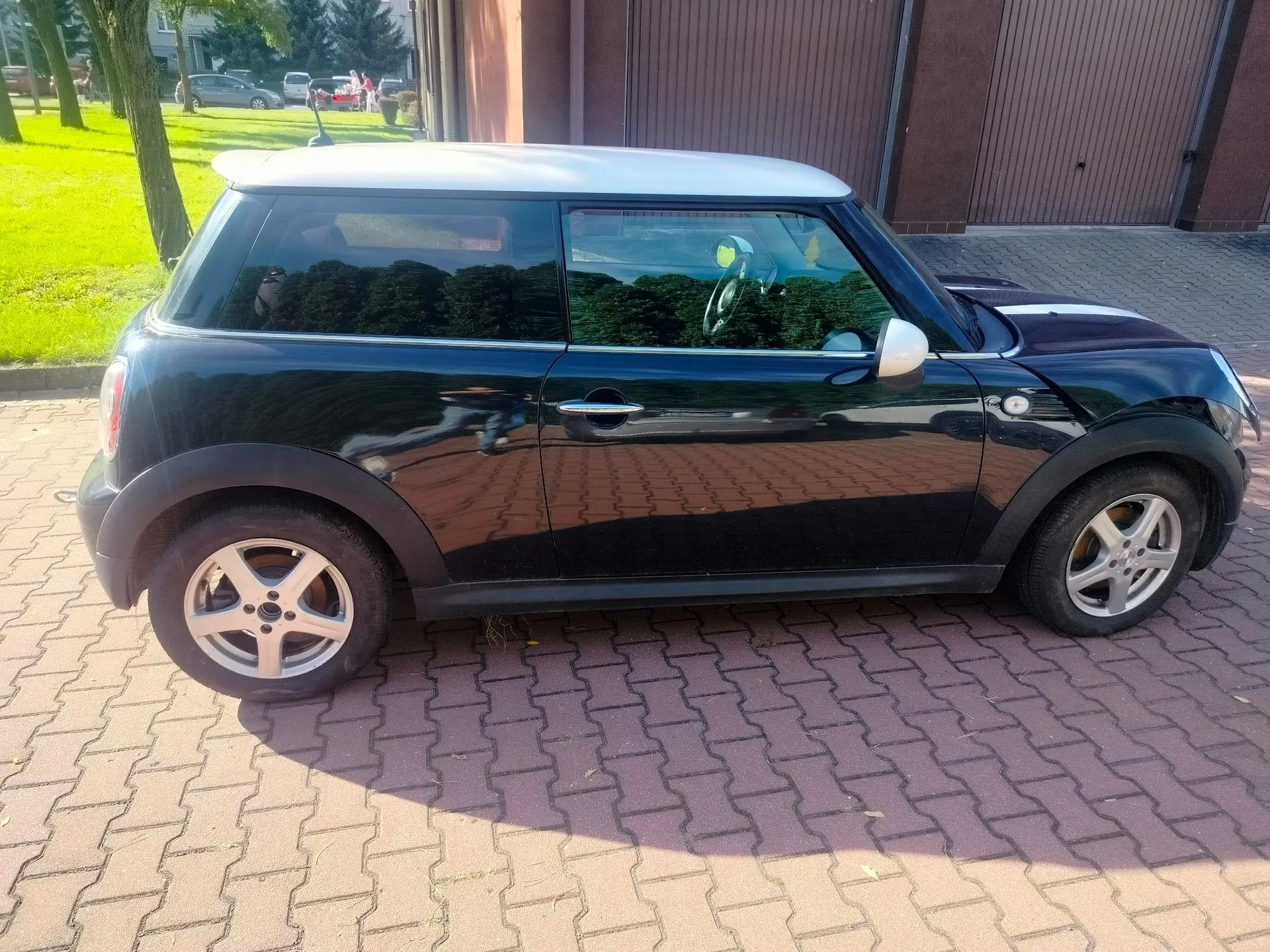 Czarny samochód Mini Cooper z białym dachem, zaparkowany bokiem na kostce brukowej. W tle widoczny jest trawnik, drzewa oraz brązowe drzwi garażowe.