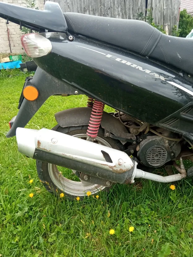 Zbliżenie na tylną część czarnego skutera marki Formax, zaparkowanego na zielonej trawie z żółtymi kwiatami. Widoczny jest srebrny tłumik wydekowy z widocznym uszkodzeniem obudowy, czerwona sprężyna amortyzatora, tylne koło oraz tylna lampa i kierunkowskaz. Na boku skutera znajduje się napis 'FORMAX'.