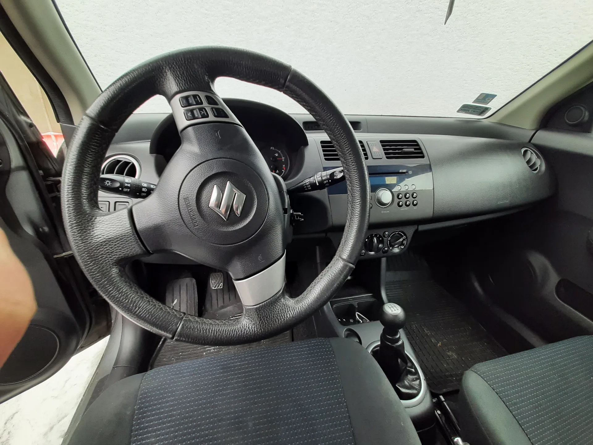 Kierownica z logo Suzuki, czarna deska rozdzielcza z przyciskami, radio, klimatyzacja, ręczna skrzynia biegów, czarne siedzenia z tkaniną, otwarte drzwi kierowcy, jasny sufit. Wnętrze w odcieniach czerni i szarości, widoczne pedały i kierunkowskazy na kierownicy.