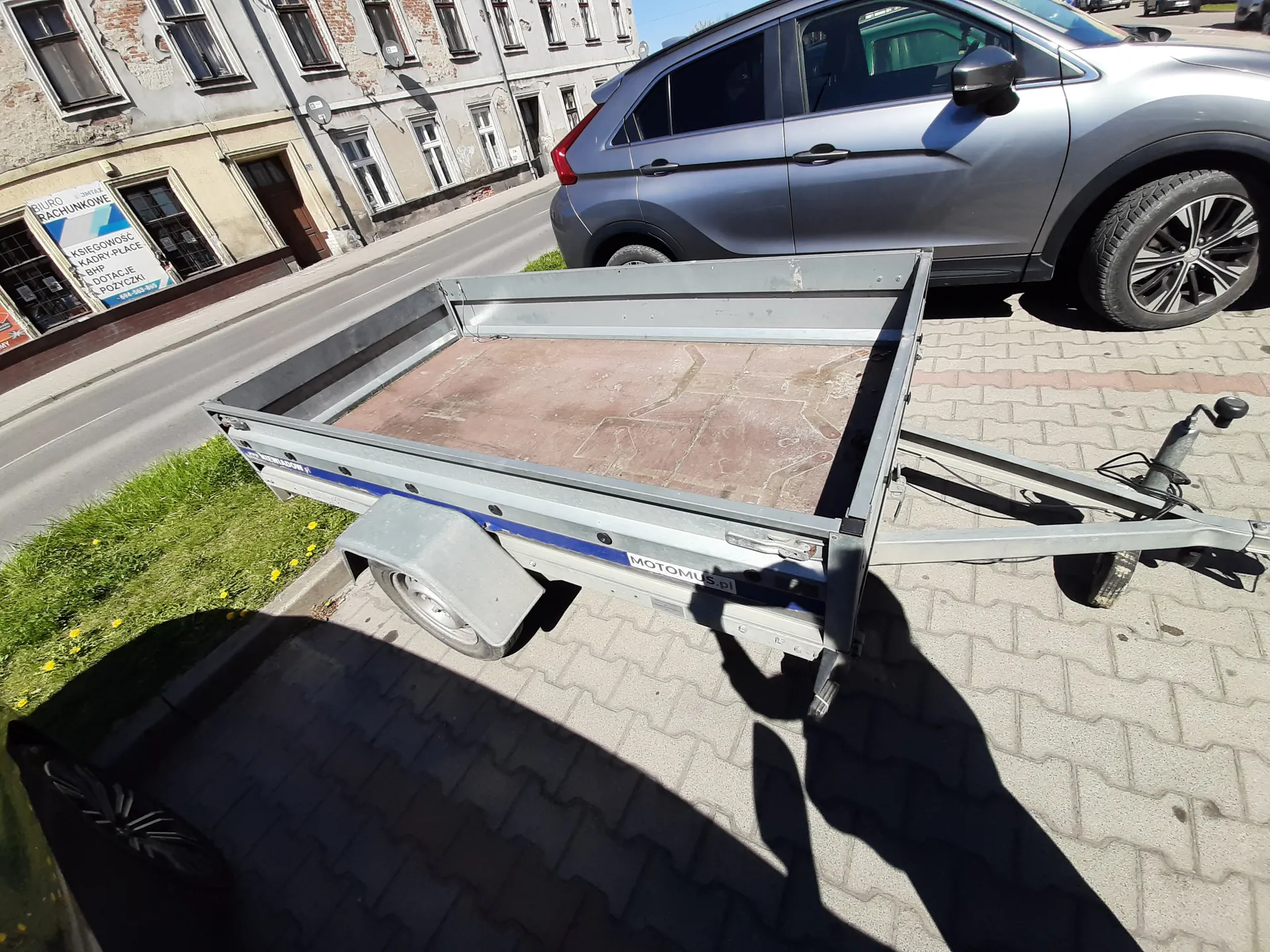 Mały przyczepa metalowa z drewnianym dnem zaparkowana na brukowanej powierzchni. Przyczepa posiada napis 'MOTOMUS.pl' na boku. Obok niej widoczny jest srebrny samochód typu SUV. W tle stoją stare budynki z widocznymi szyldami: 'BIURO RACHUNKOWE', 'KSIĘGOWOŚĆ', 'KADRY-PLACE', 'BHP', 'POŻYCZKI'. Na lewo od przyczepy znajduje się trawnik z żółtymi kwiatami. Scena rozgrywa się na zewnątrz w słoneczny dzień.