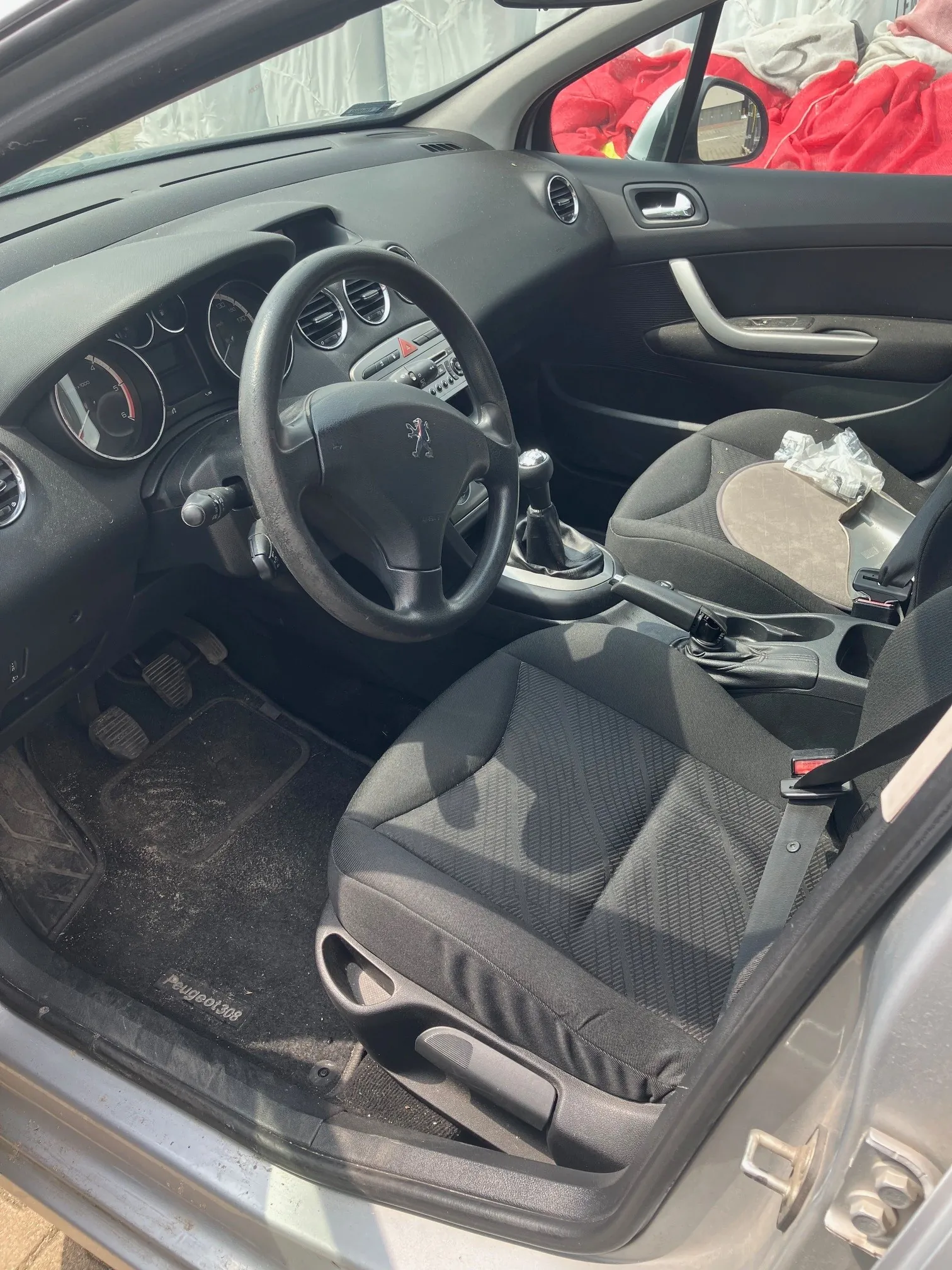 Wnętrze samochodu Peugeot 308 widziane z otwartych drzwi kierowcy. Widoczna jest czarna tapicerka foteli, kierownica, deska rozdzielcza z zegarami oraz pedały. Na dywaniku w podłodze widnieje napis 'Peugeot 308'. W tle, za szybą, widać czerwony materiał.