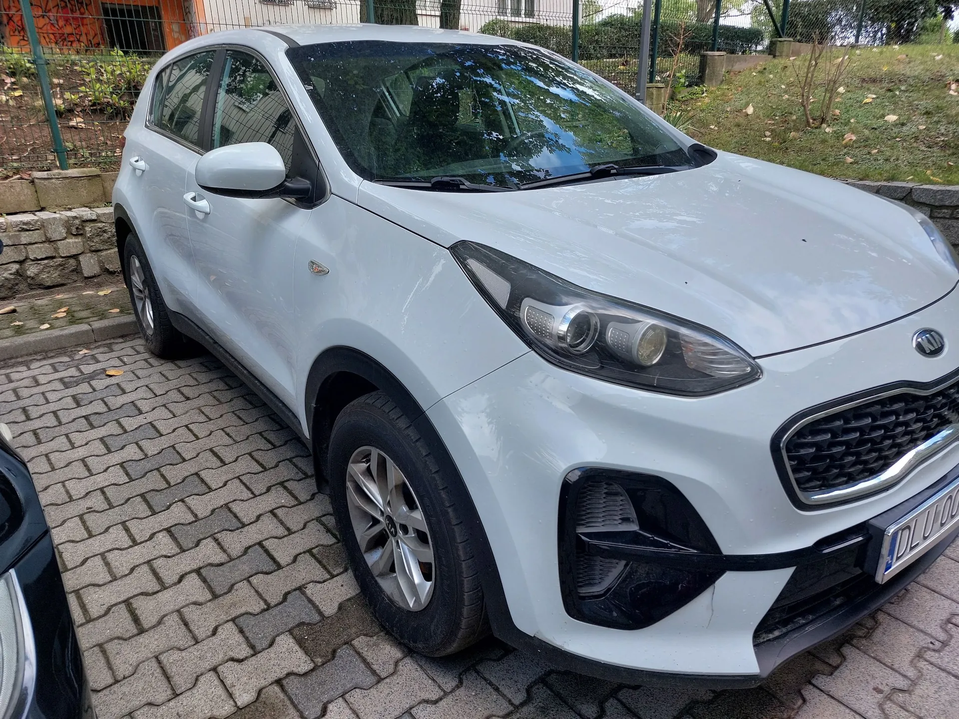 Biały samochód marki Kia typu SUV zaparkowany na kostce brukowej. Pojazd jest widoczny z przodu i z boku, z perspektywy trzech czwartych. W tle widać zielone krzewy, ogrodzenie z siatki oraz fragmenty trawnika.